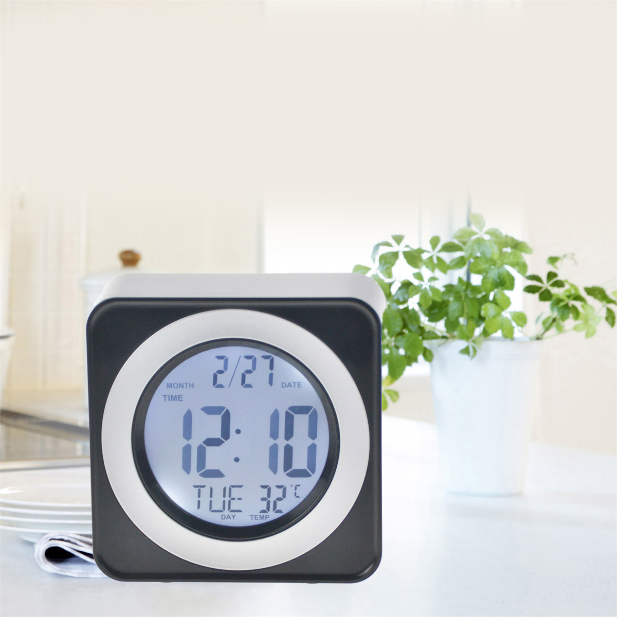 Digitaluhr zeigt 12:10, Di 27 und 32 Grad an. Auf weißem Tisch neben Fenster und Pflanze.