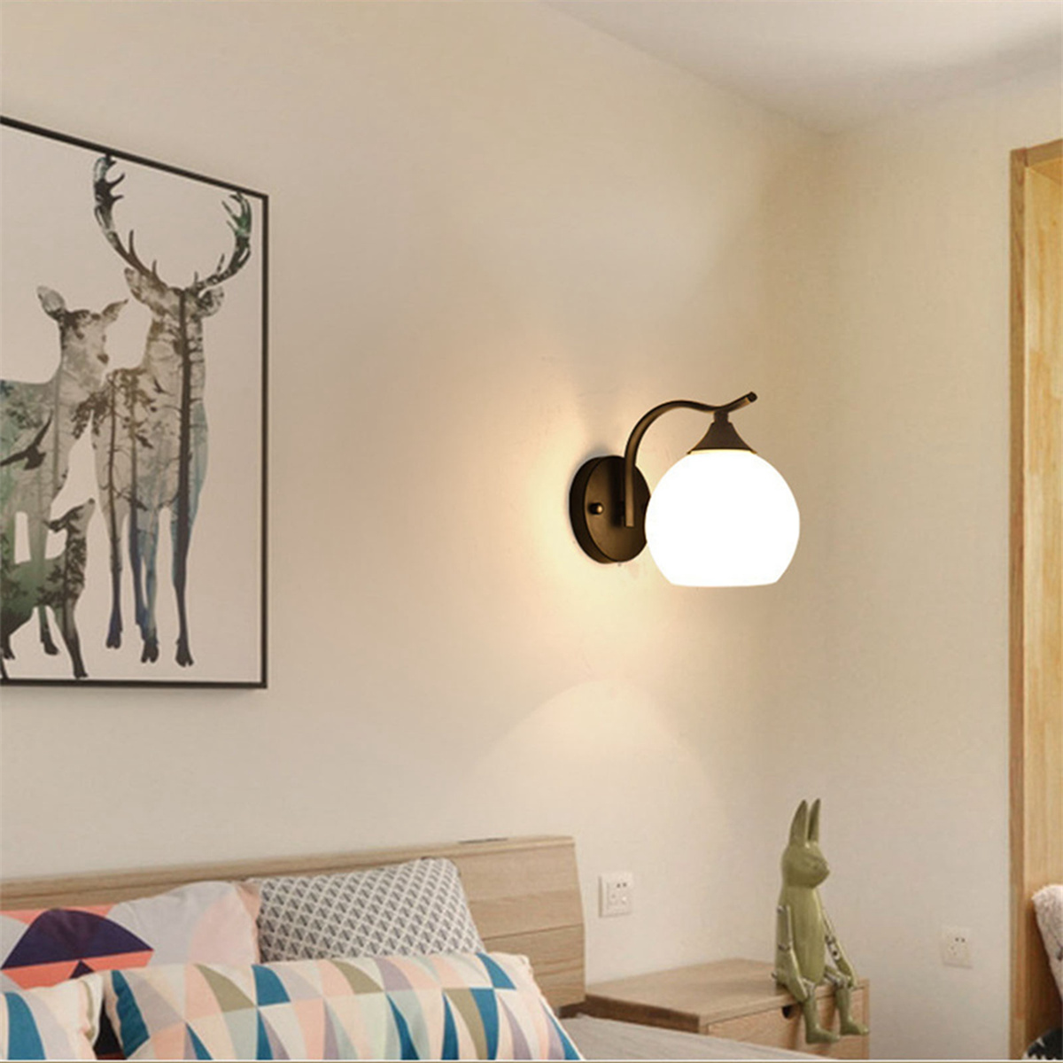 Eine Wandlampe mit weißer Glühbirne und ein Gemälde von Hirschen. Ein kleines dekoratives Kaninchen sitzt daneben.