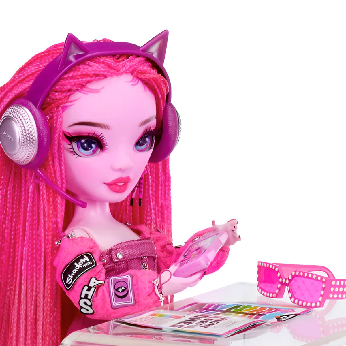 Różowa lalka ze słuchawkami, okularami i magazynem na biurku. Różowy motyw dominuje.