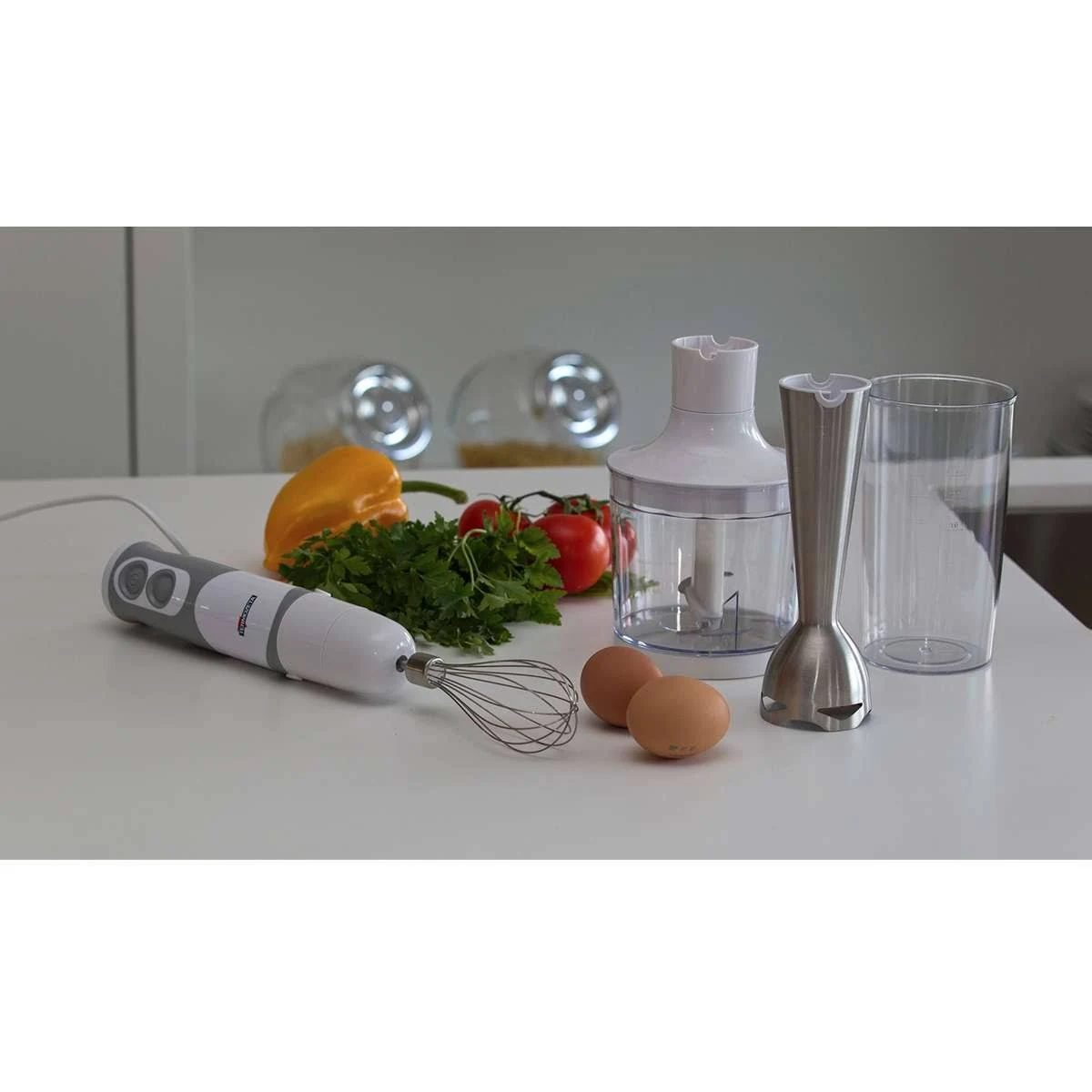 Frullatore e accessori su un tavolo bianco. Include uova, pomodori, peperoni, prezzemolo. Ambiente luminoso.