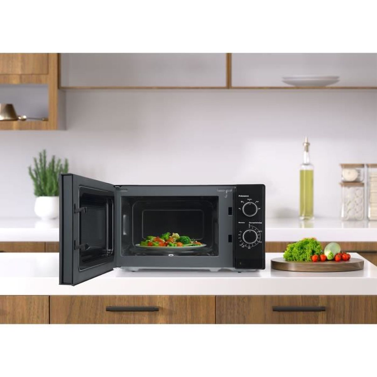 Microondas negro con puerta abierta, plato de comida dentro y controles. Verduras en el mostrador.