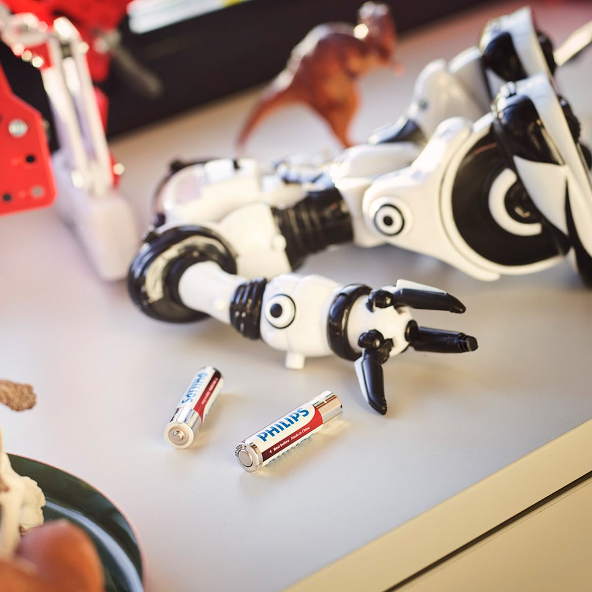 Zabawki na stole: robot, dinozaur i baterie. Biała powierzchnia i rozmyte tło.