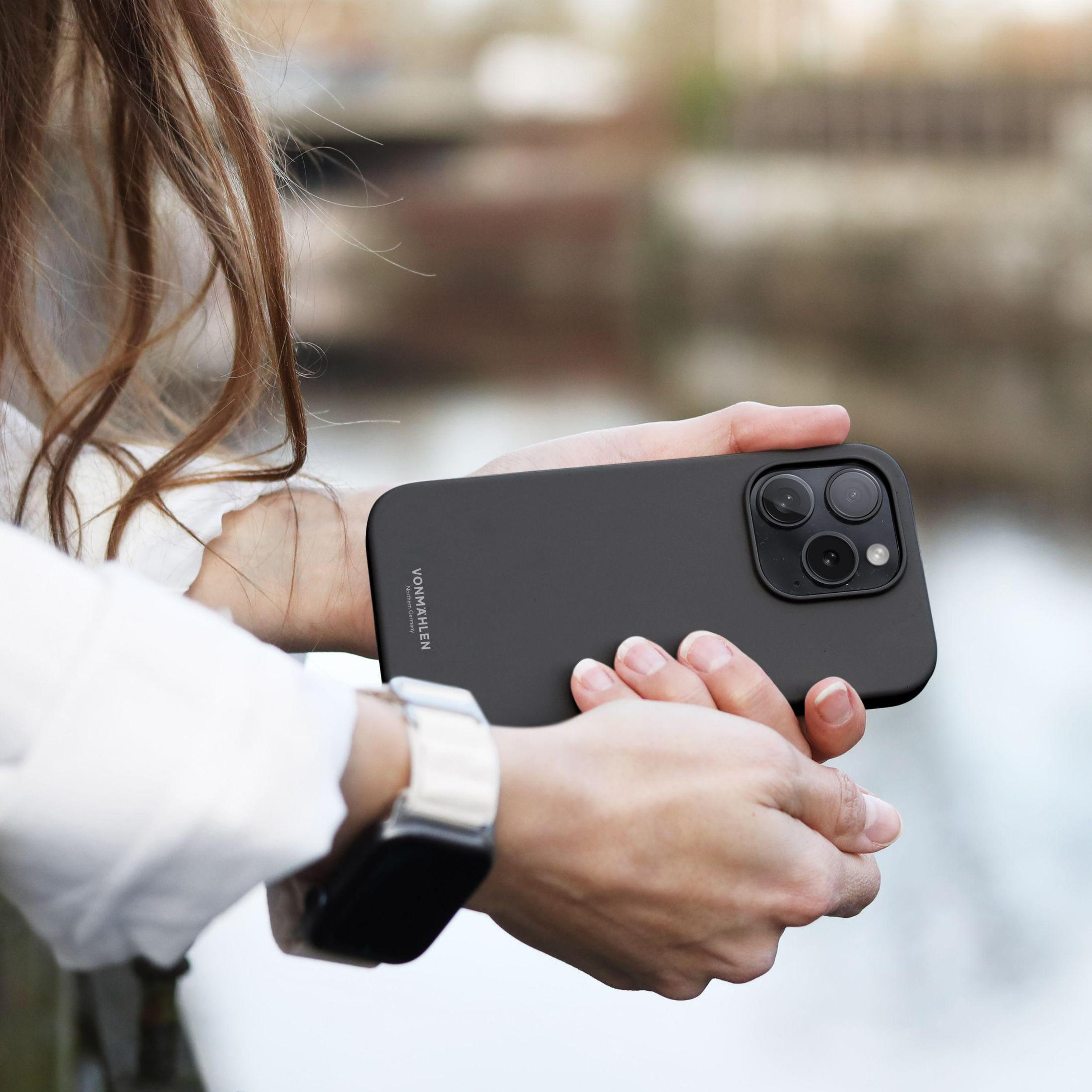 Persona che tiene un telefono nero con tripla fotocamera, orologio e sfondo sfocato.