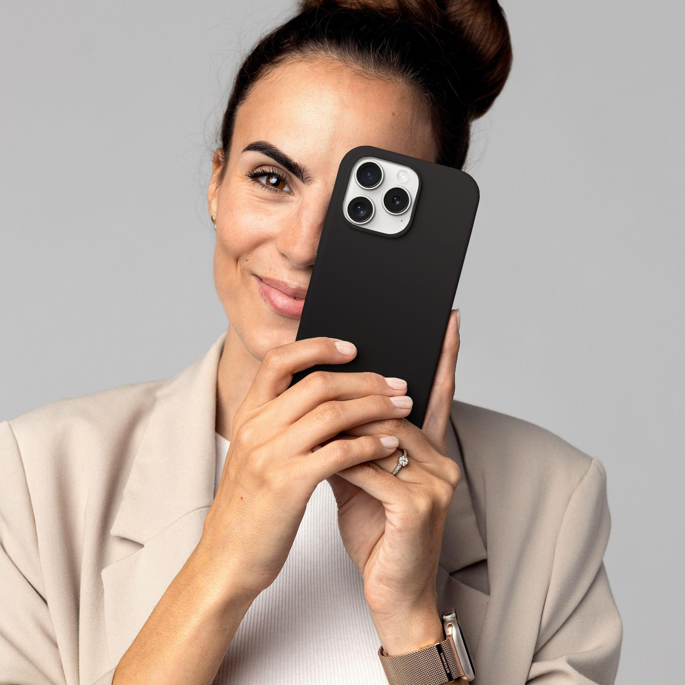 Eine Frau, die ein schwarzes Telefon vor ihrem Gesicht hält, lächelt, trägt Blazer und Uhr.