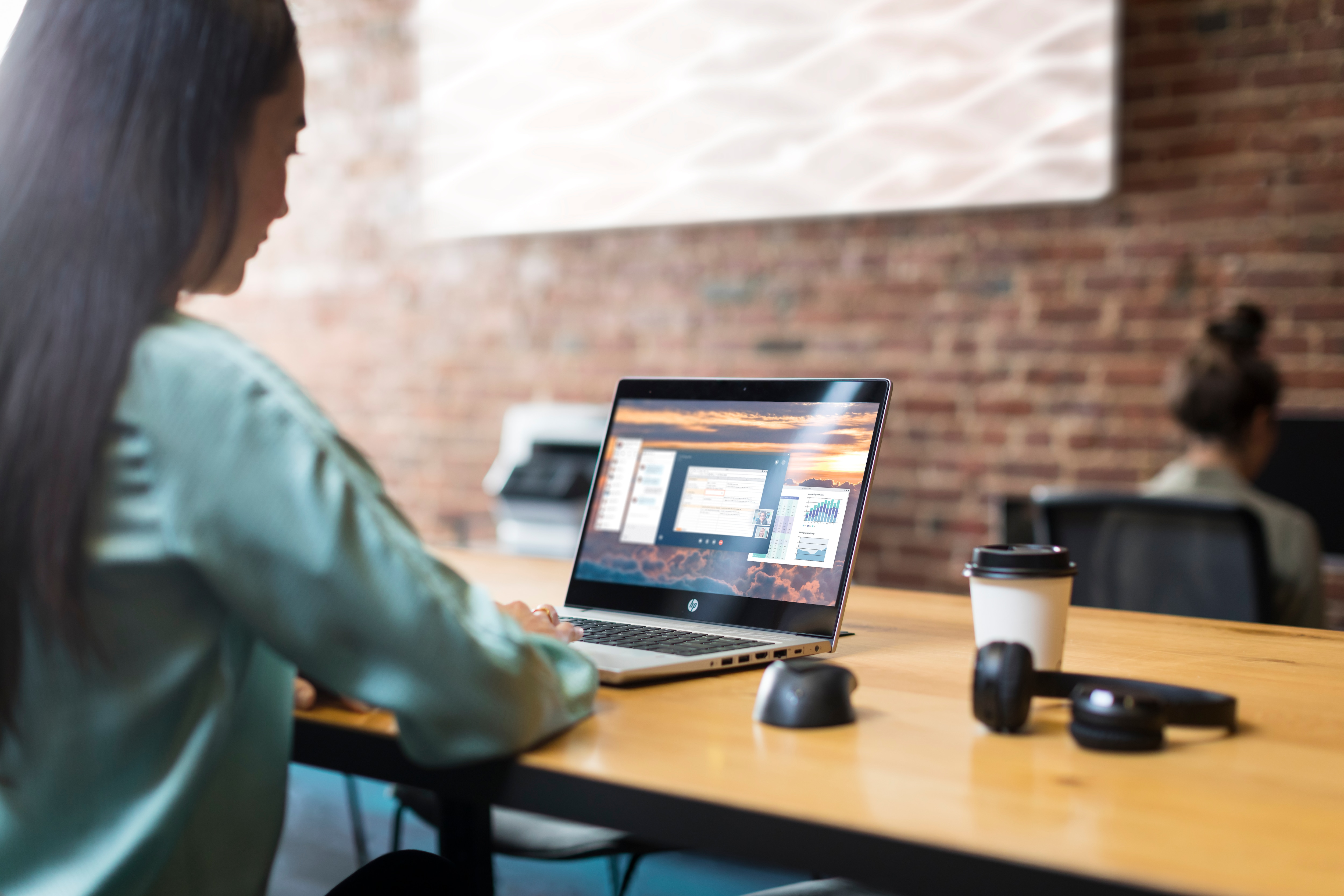 Kobieta pracuje na laptopie przy drewnianym biurku. Są słuchawki i filiżanka kawy.