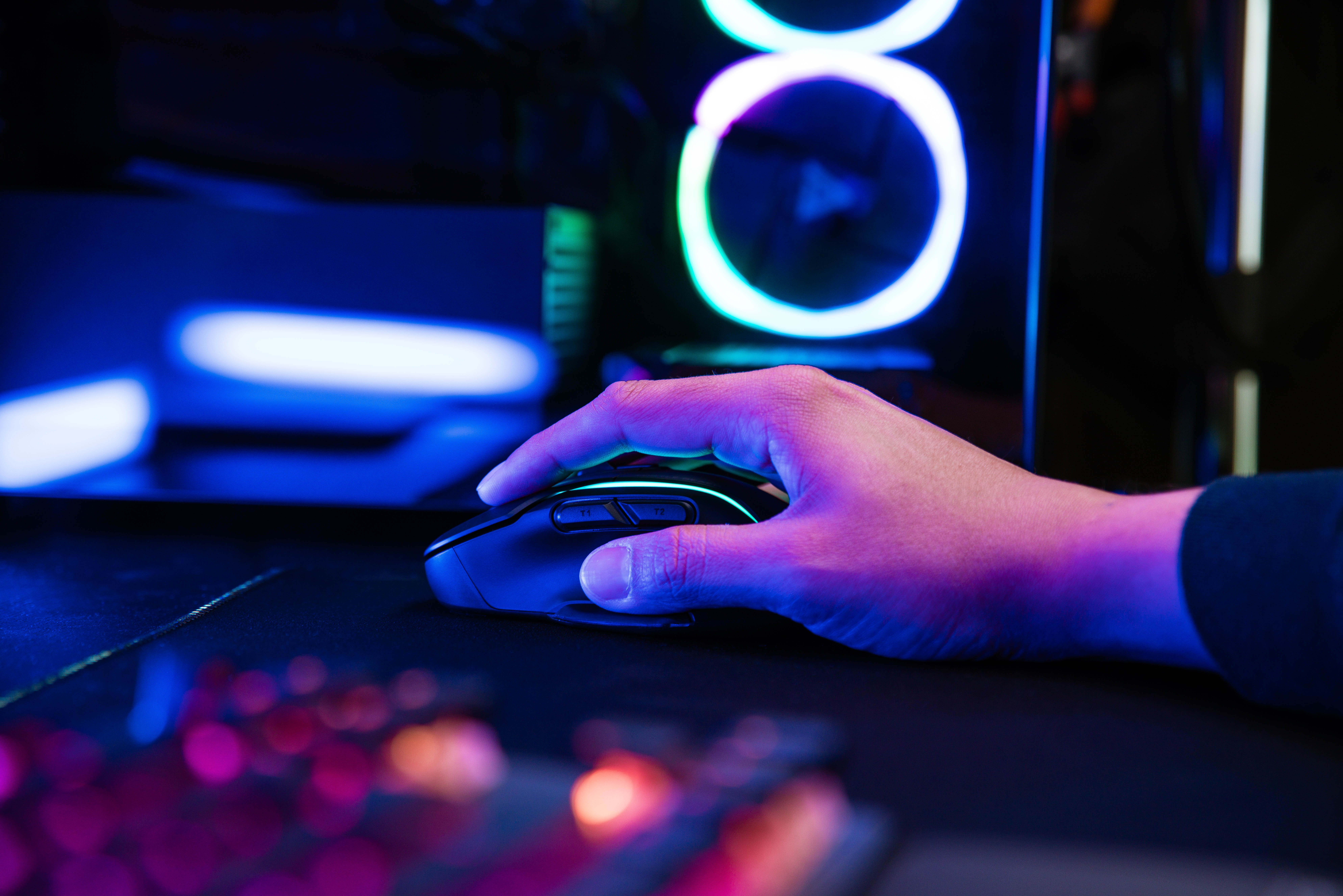 Dłoń używająca myszy gamingowej na czarnej podkładce w pobliżu klawiatury i komputera.