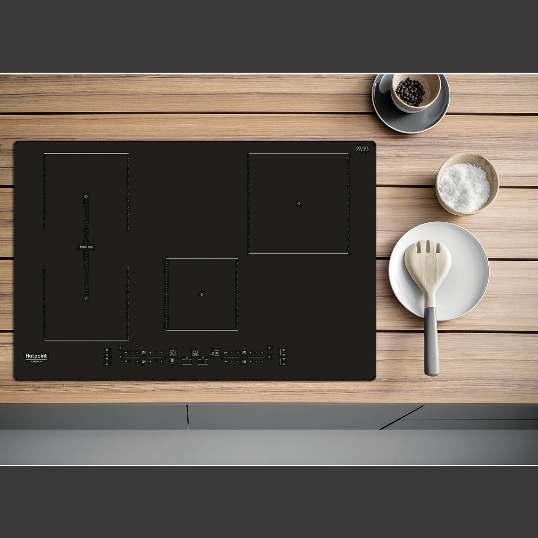 Czarna płyta grzejna z czterema palnikami. Biały talerz, miska z solą i łyżka na drewnianym blacie.