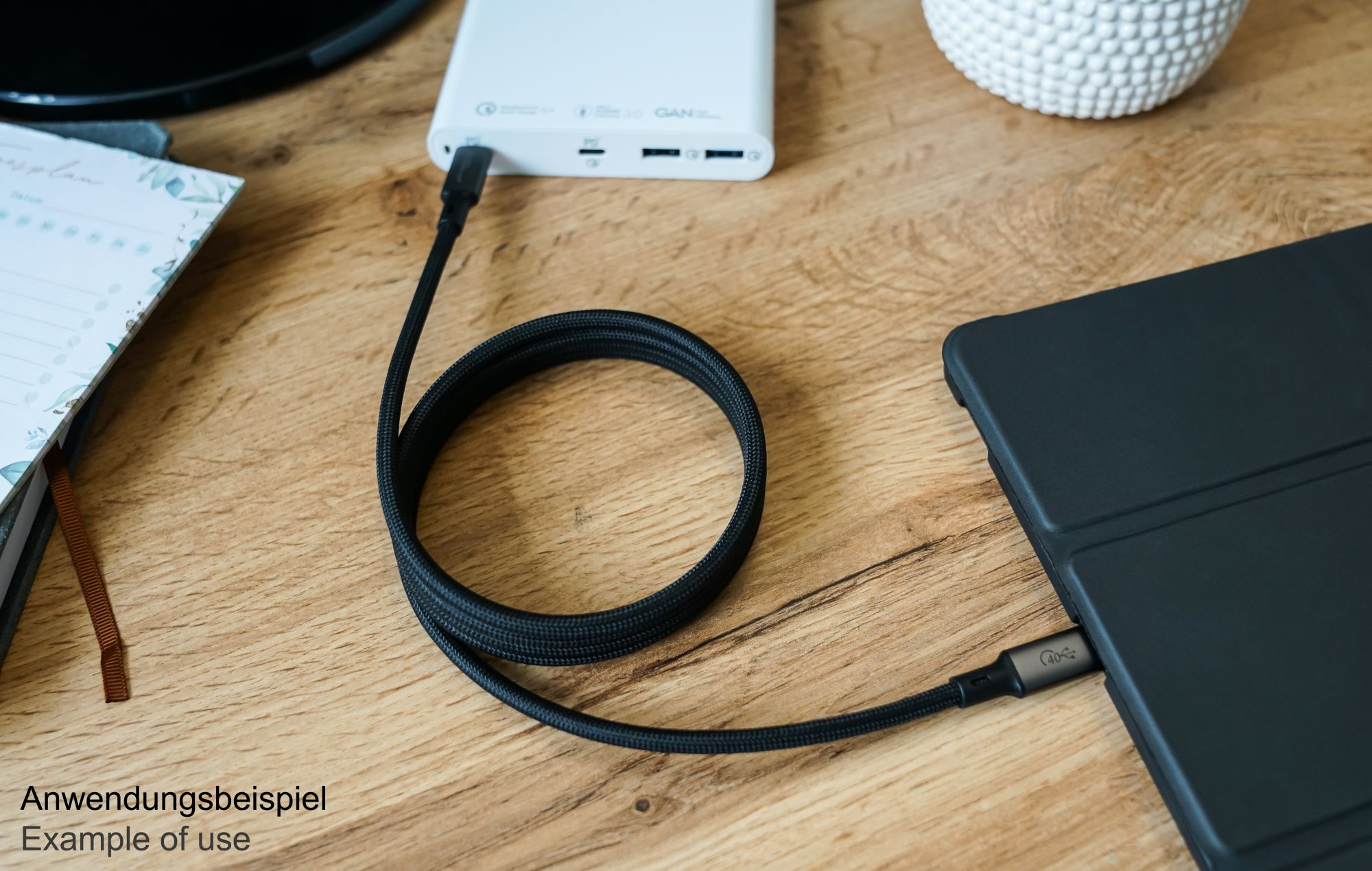 Schwarzes USB-Kabel, verbunden mit einer weißen Powerbank und einem schwarzen Tablet. Text unten: Anwendungsbeispiel.