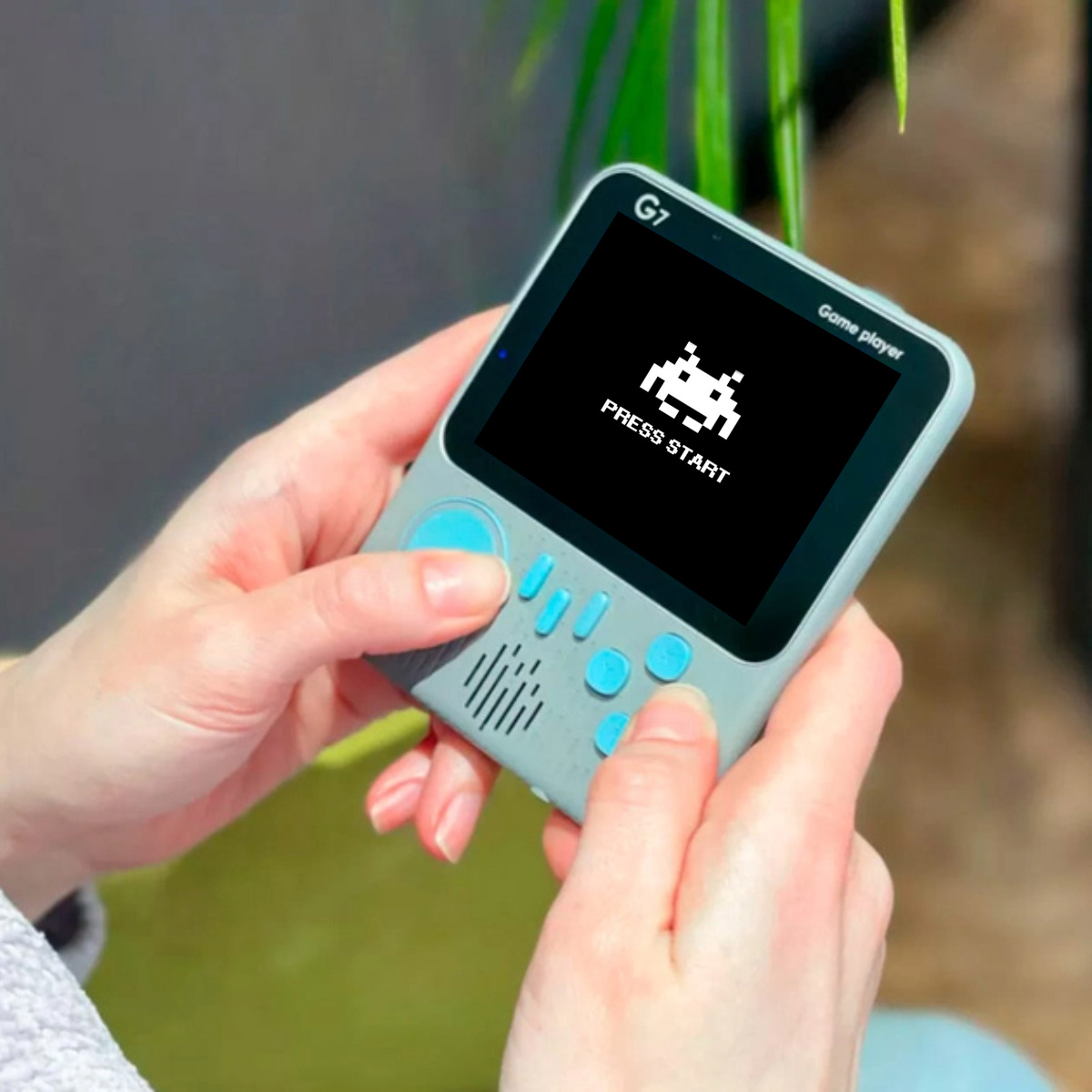 Consola retro portátil mostrando un juego. Muestra un alien pixelado y 'Press Start'.