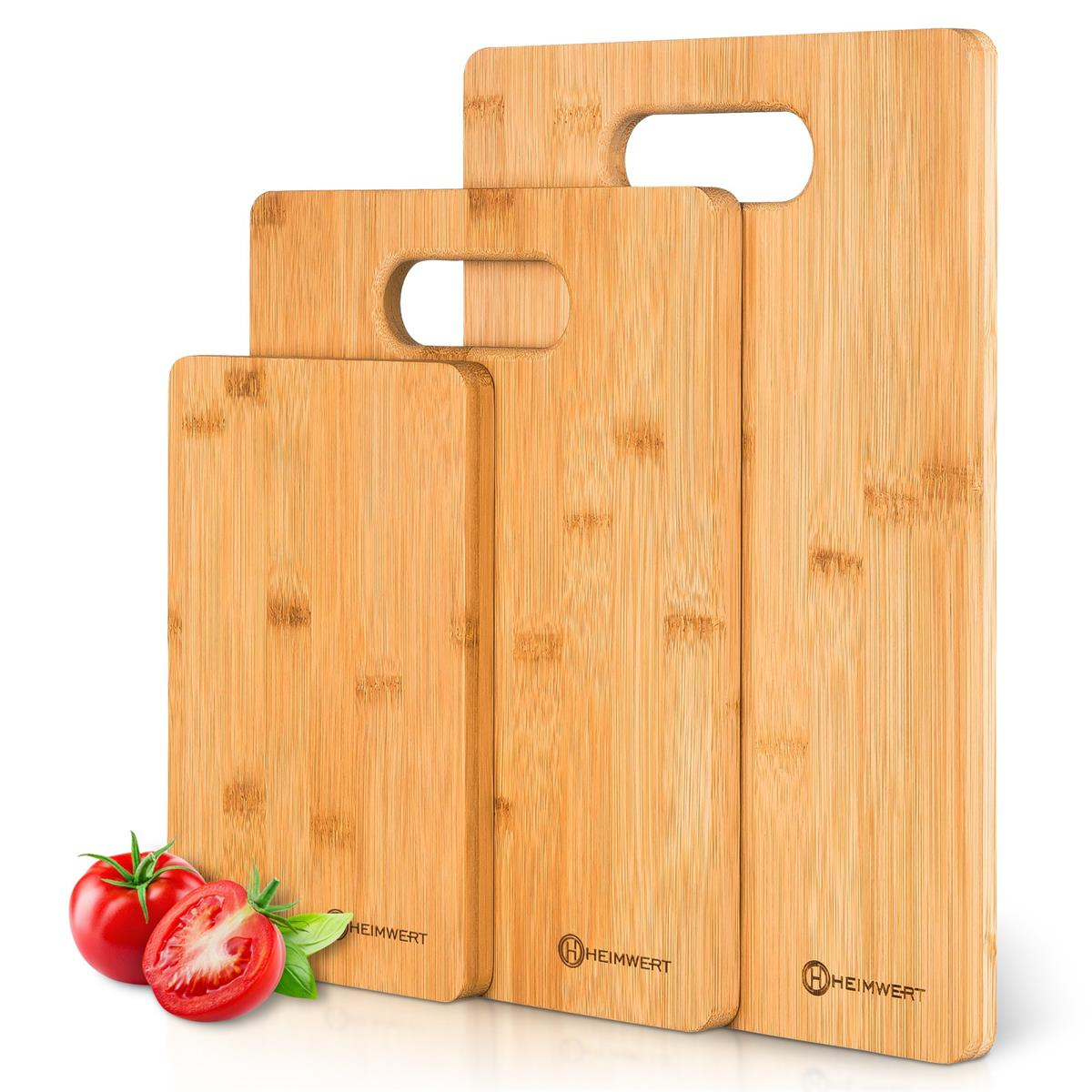 Drei Holzschneidebretter mit Griffen. Zwei Tomaten und Basilikumblätter sind links.