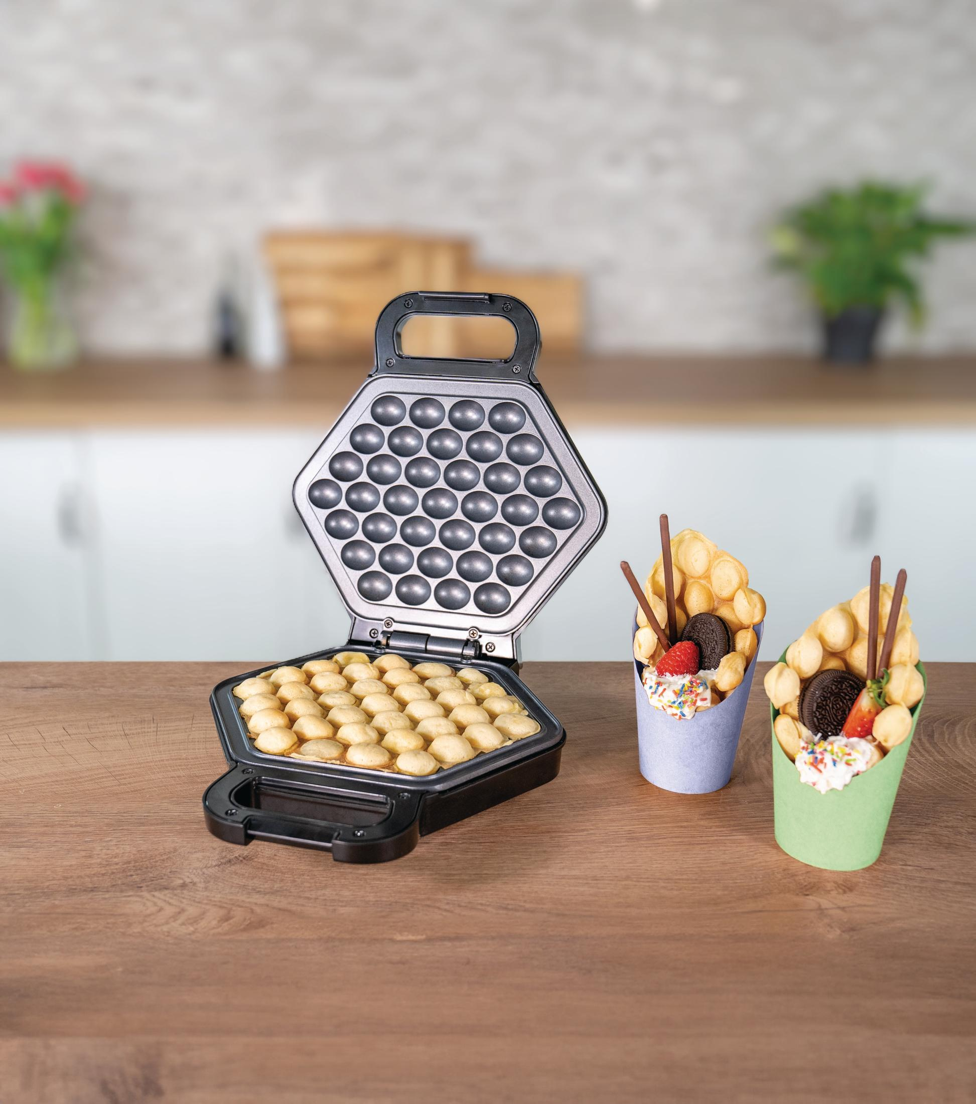 Otwarta gofrownica z upieczonymi goframi i dwoma rożkami do gofrów z dodatkami na drewnianym stole.