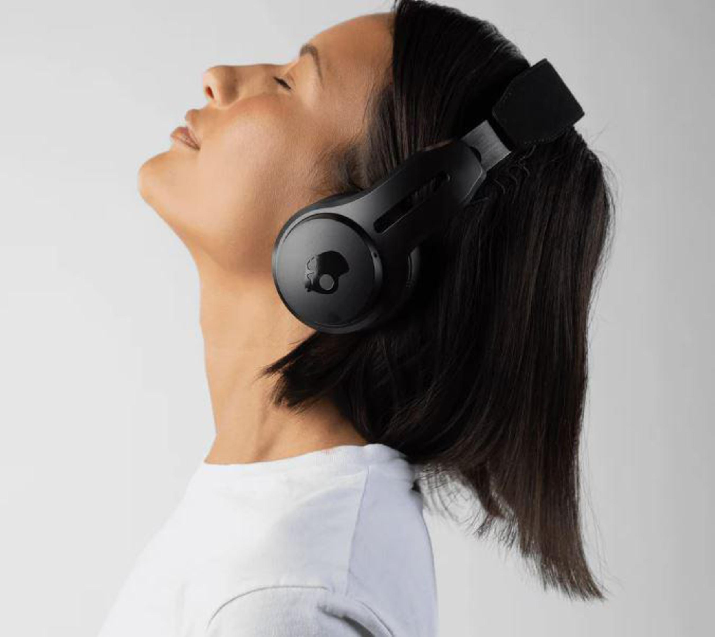 Kobieta ze słuchawkami na głowie, patrząca w górę. Czarne słuchawki, biała koszulka.