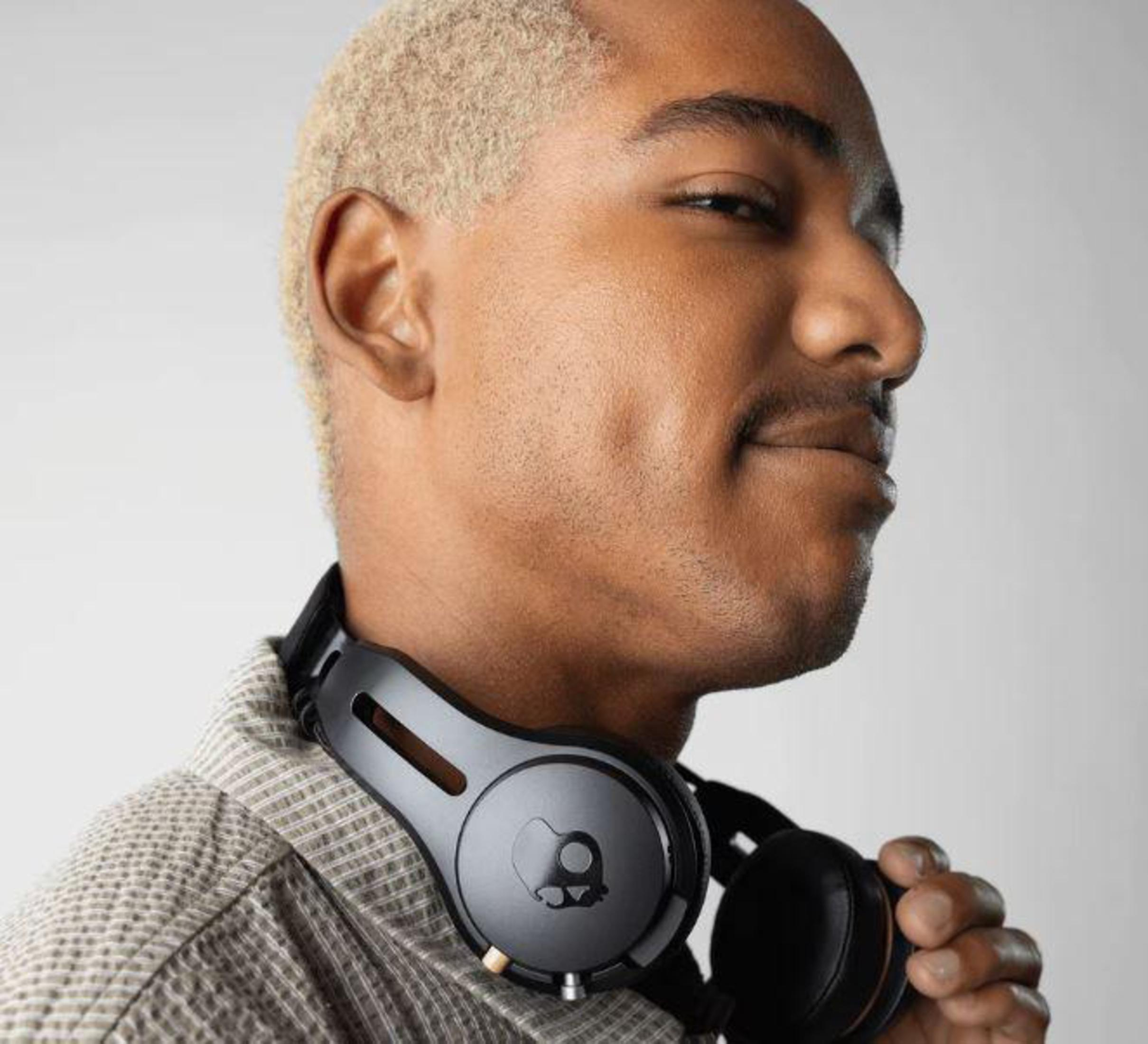 Mężczyzna o jasnej karnacji z blond włosami noszący czarne słuchawki. Ma małe wąsy i dołeczek.