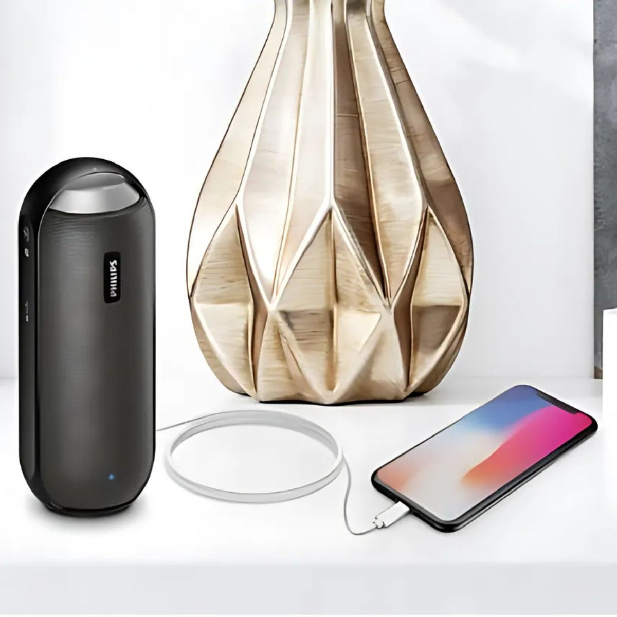 Czarny głośnik Philips, złoty wazon, biały kabel i smartfon ładujący się na białej powierzchni. Neutralne tło.