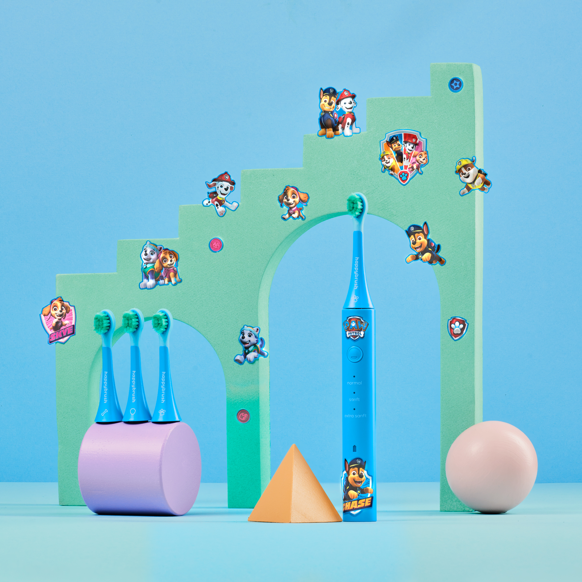 Cepillo de dientes eléctrico con personajes de Paw Patrol, accesorios y una esfera rosa sobre fondo azul.