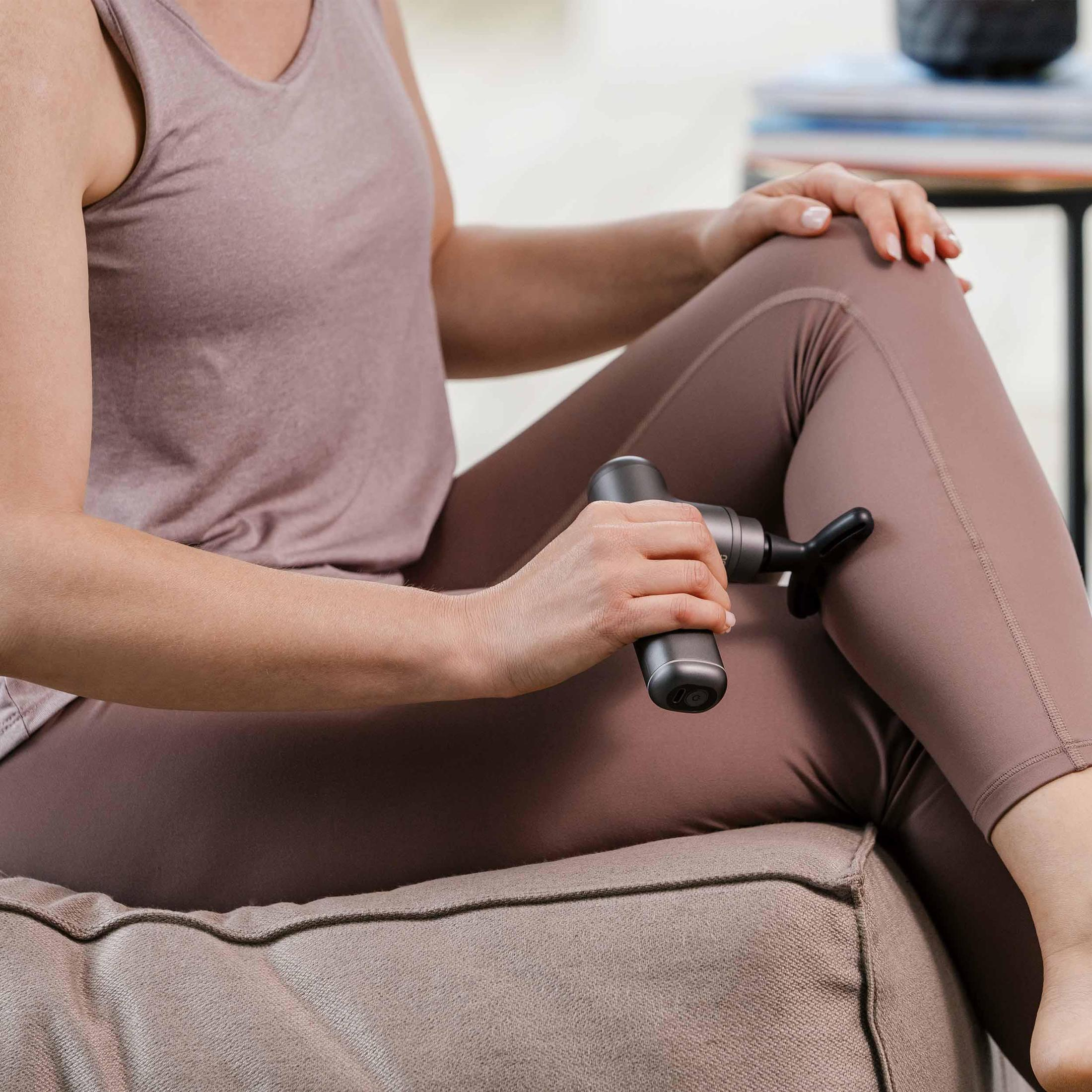 Frau benutzt ein Massagegerät an ihrem Bein im Sitzen. Sie trägt ein Tanktop und Leggings.