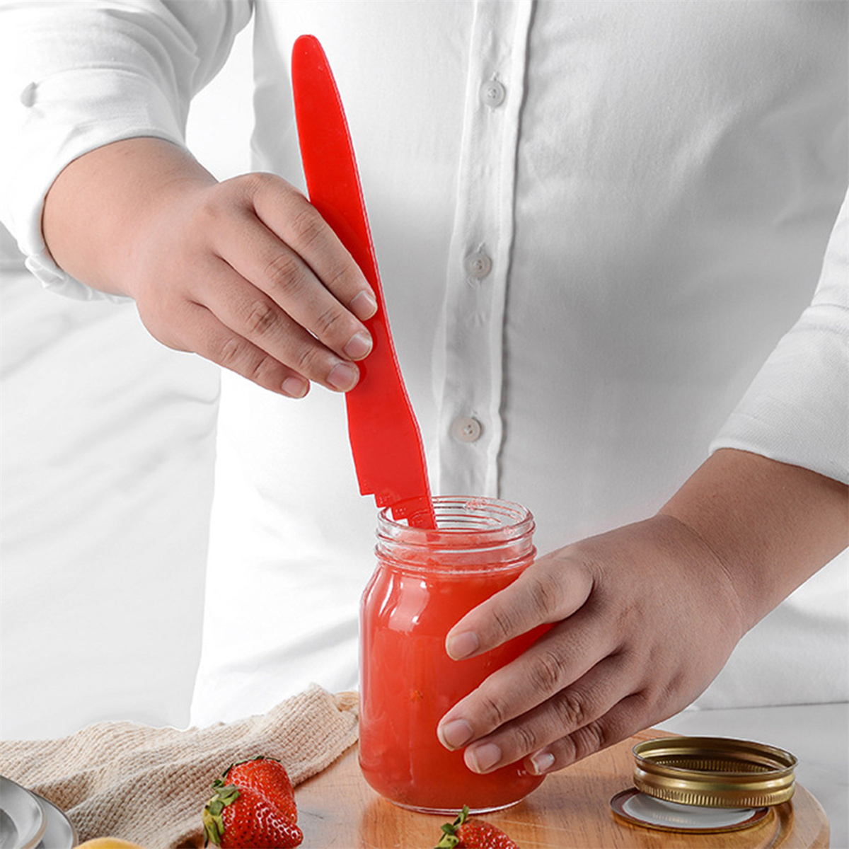 Person öffnet mit rotem Werkzeug ein Glas mit roter Flüssigkeit. Erdbeeren und Deckel liegen daneben.