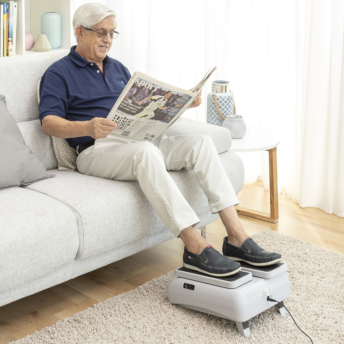 Ein älterer Mann liest Zeitung, während er ein Fußmassagegerät benutzt. Er sitzt auf einem grauen Sofa.