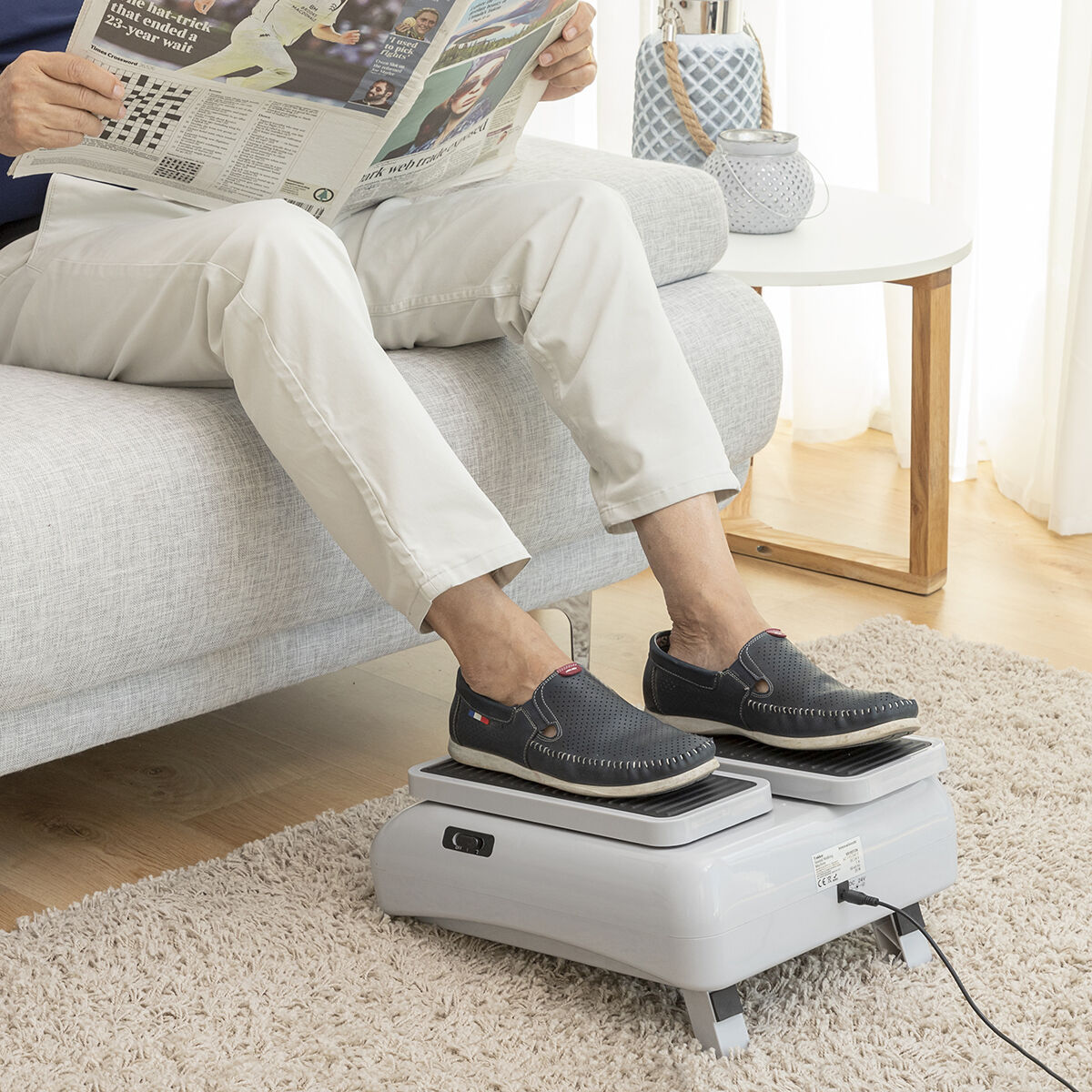 Eine Person sitzt auf einem Sofa, liest Zeitung, Füße auf einem Fußmassagegerät.
