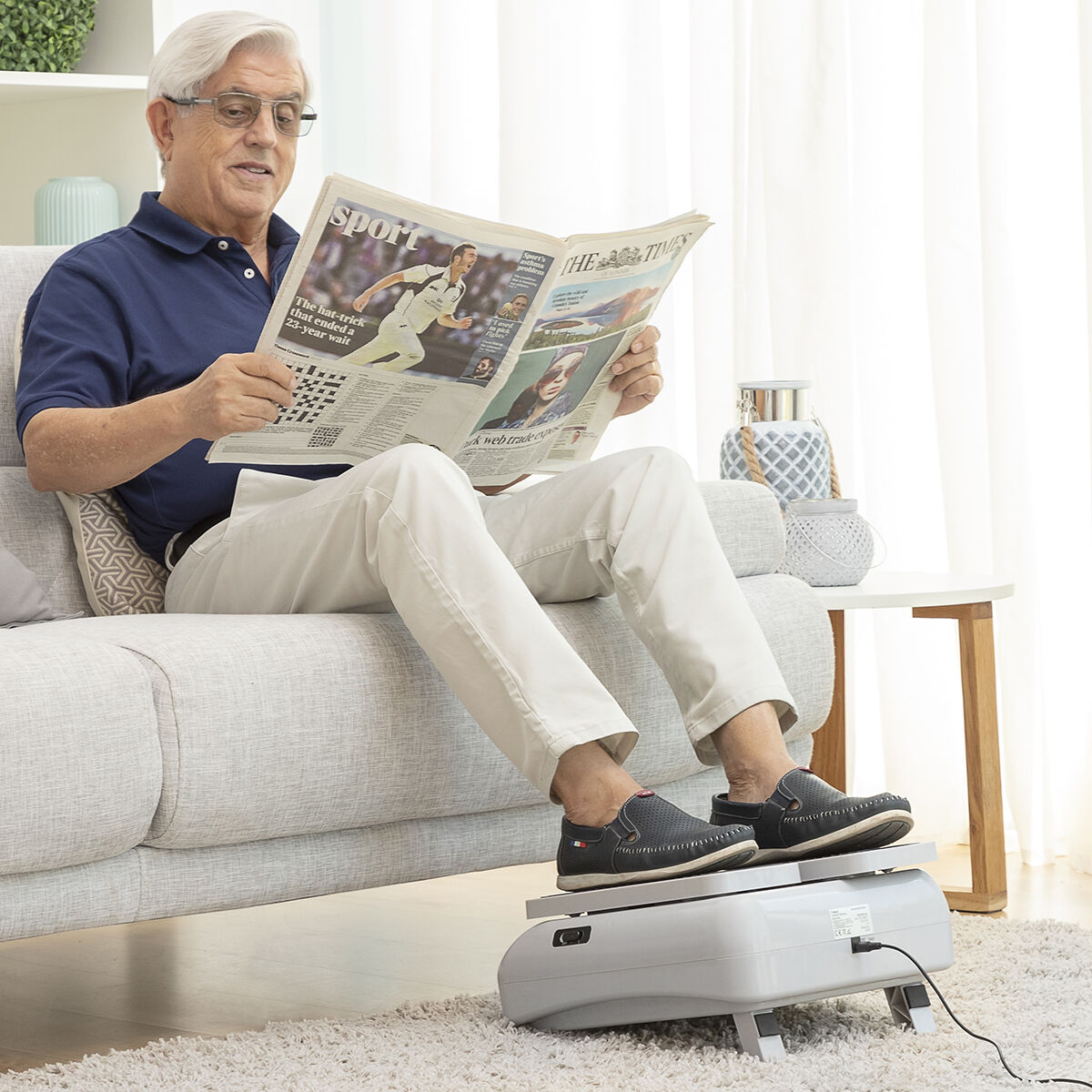 Ein älterer Mann liest eine Zeitung, während er auf einem Sofa ein Fußmassagegerät benutzt.