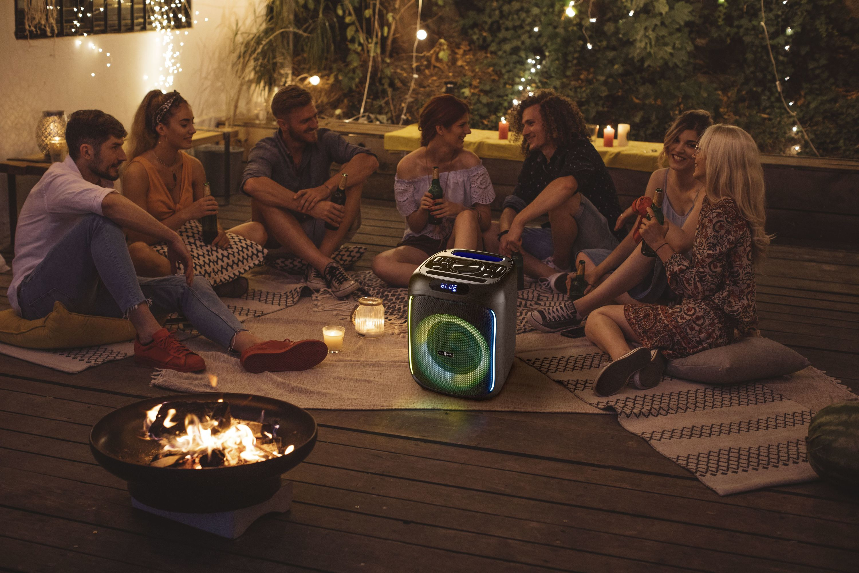 Grupa osób siedzi w nocy wokół głośnika i paleniska.