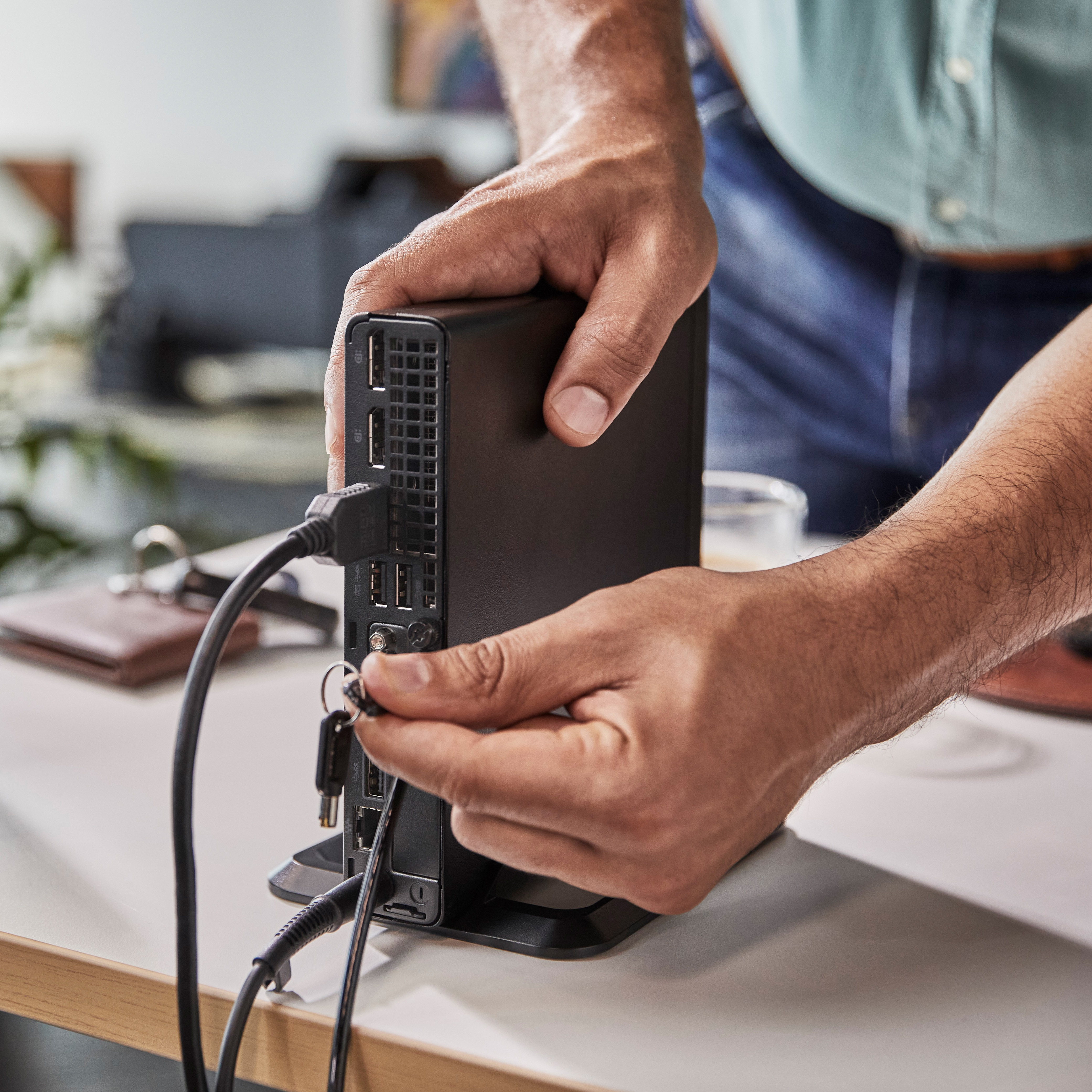 Ręce podłączają kable do czarnego urządzenia na białym biurku. Tło obejmuje biurko i artykuły biurowe.