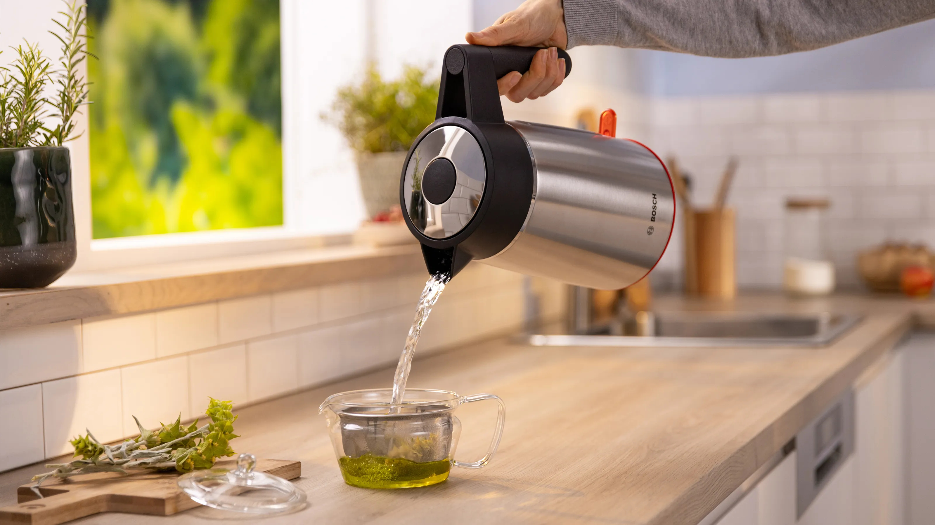 A person pouring water from a red and silver kettle into a glass teapot with tea.