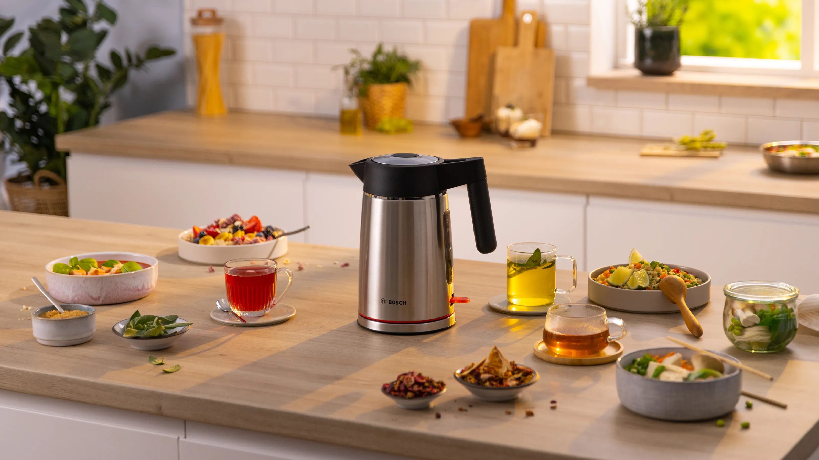A stainless steel kettle on a wooden table, surrounded by food and drinks.