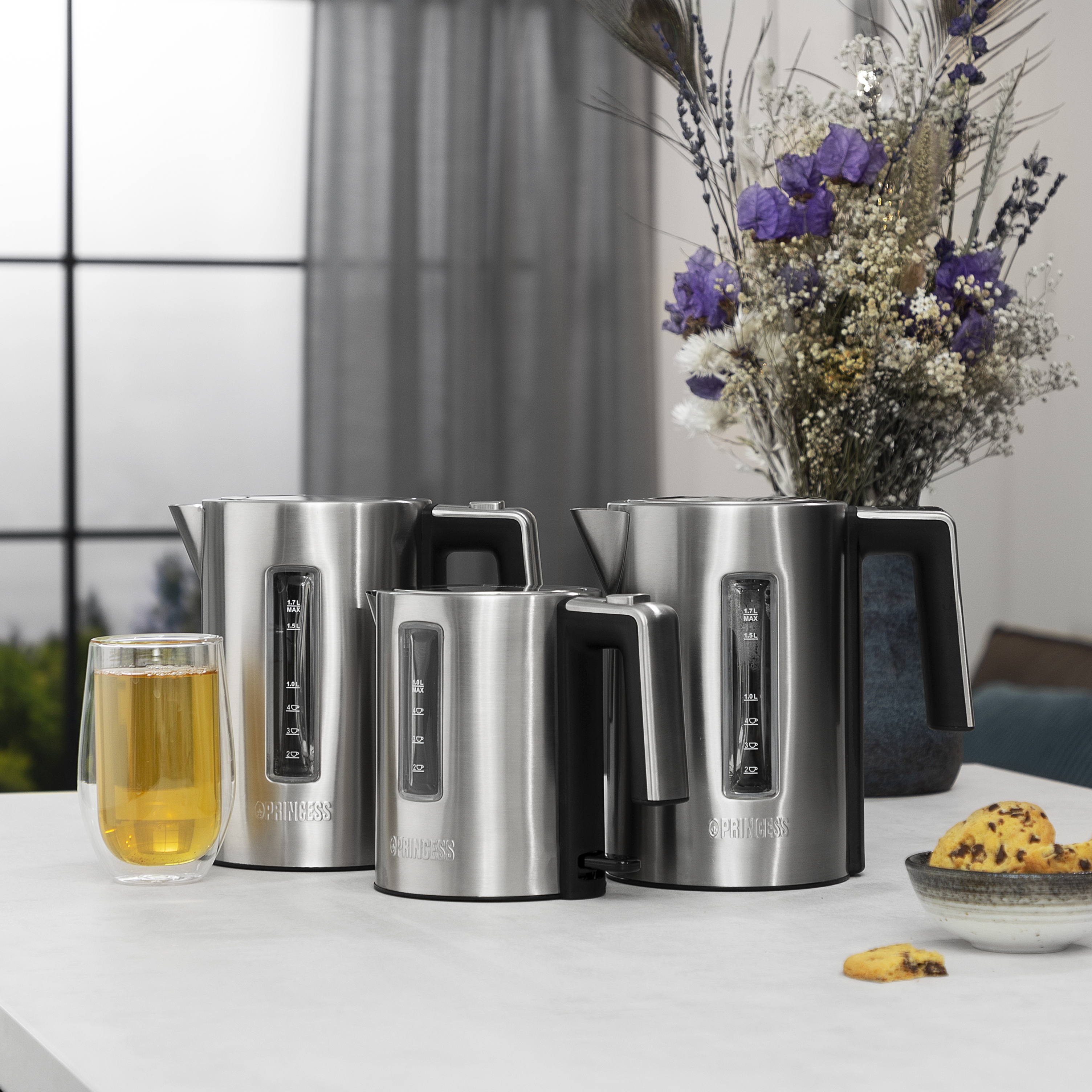 Trzy czajniki ze stali nierdzewnej o różnych rozmiarach, obok szklanki herbaty i miski ciasteczek. Kwiaty w tle.