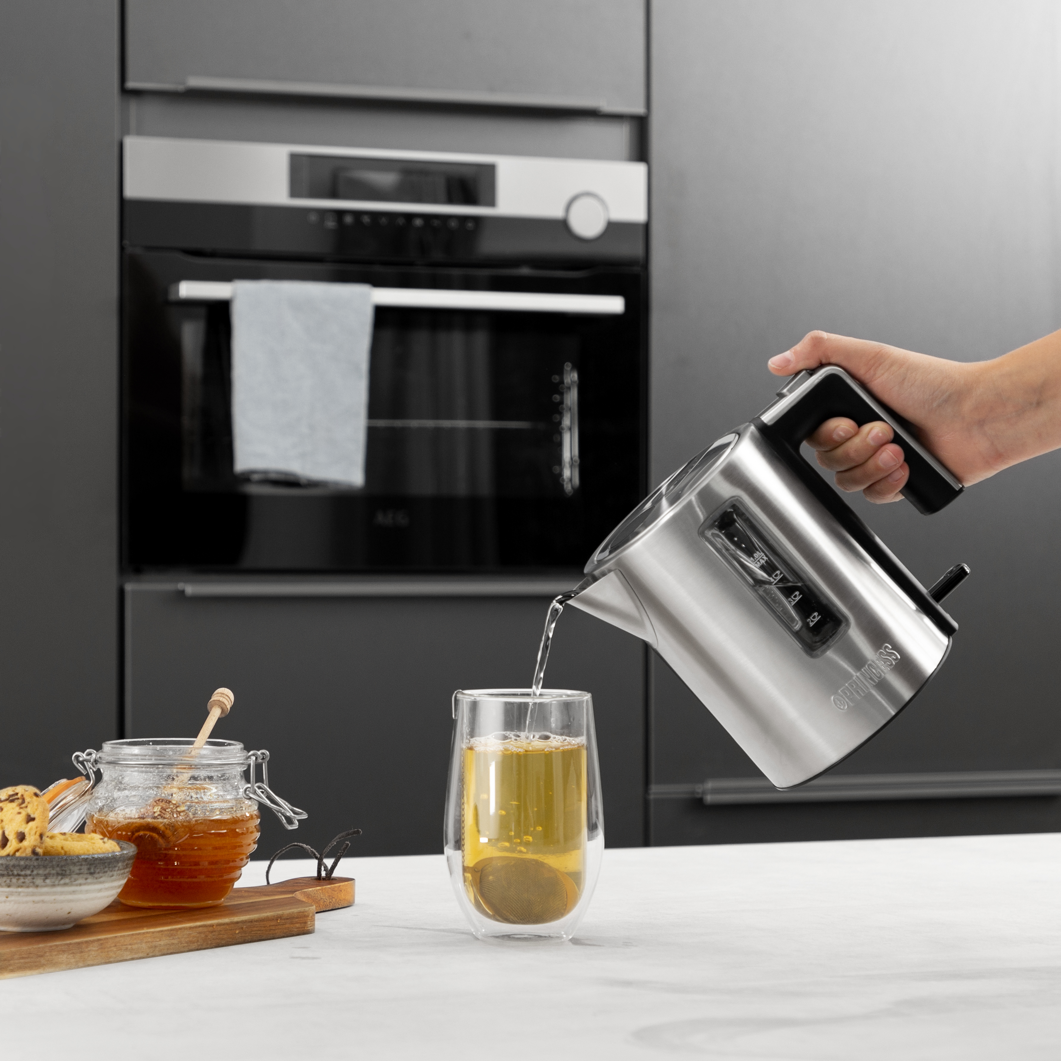 Ręka nalewająca herbatę ze stalowego czajnika do szklanki. Miód i ciastka są na stole.