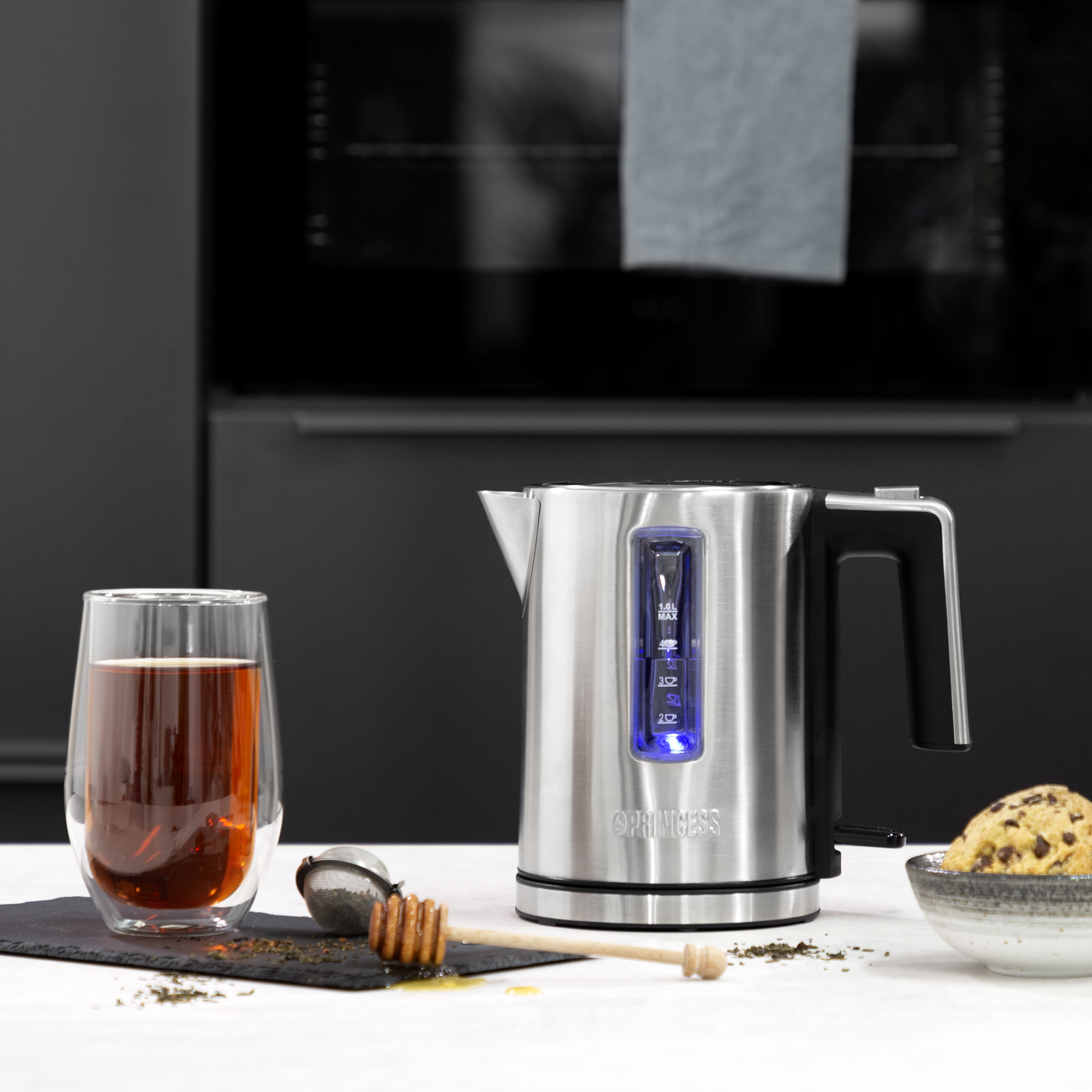 Elektryczny czajnik Princess ze stali nierdzewnej z niebieskim podświetleniem. W pobliżu szklanka herbaty i miska ciasteczek.