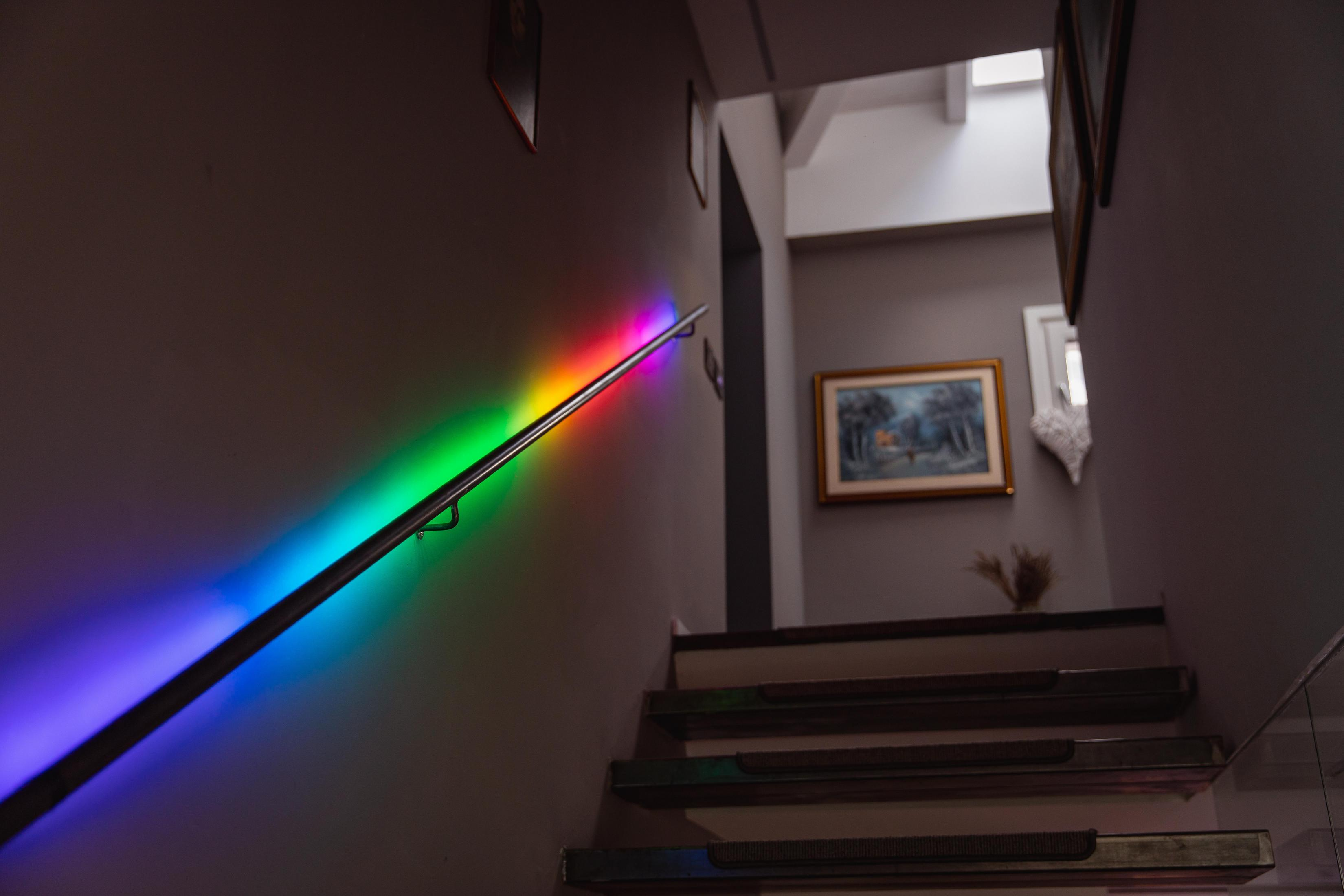 Schody z tęczowym oświetleniem LED na poręczy. Widoczne stopnie. Szare ściany i obraz na ścianie.