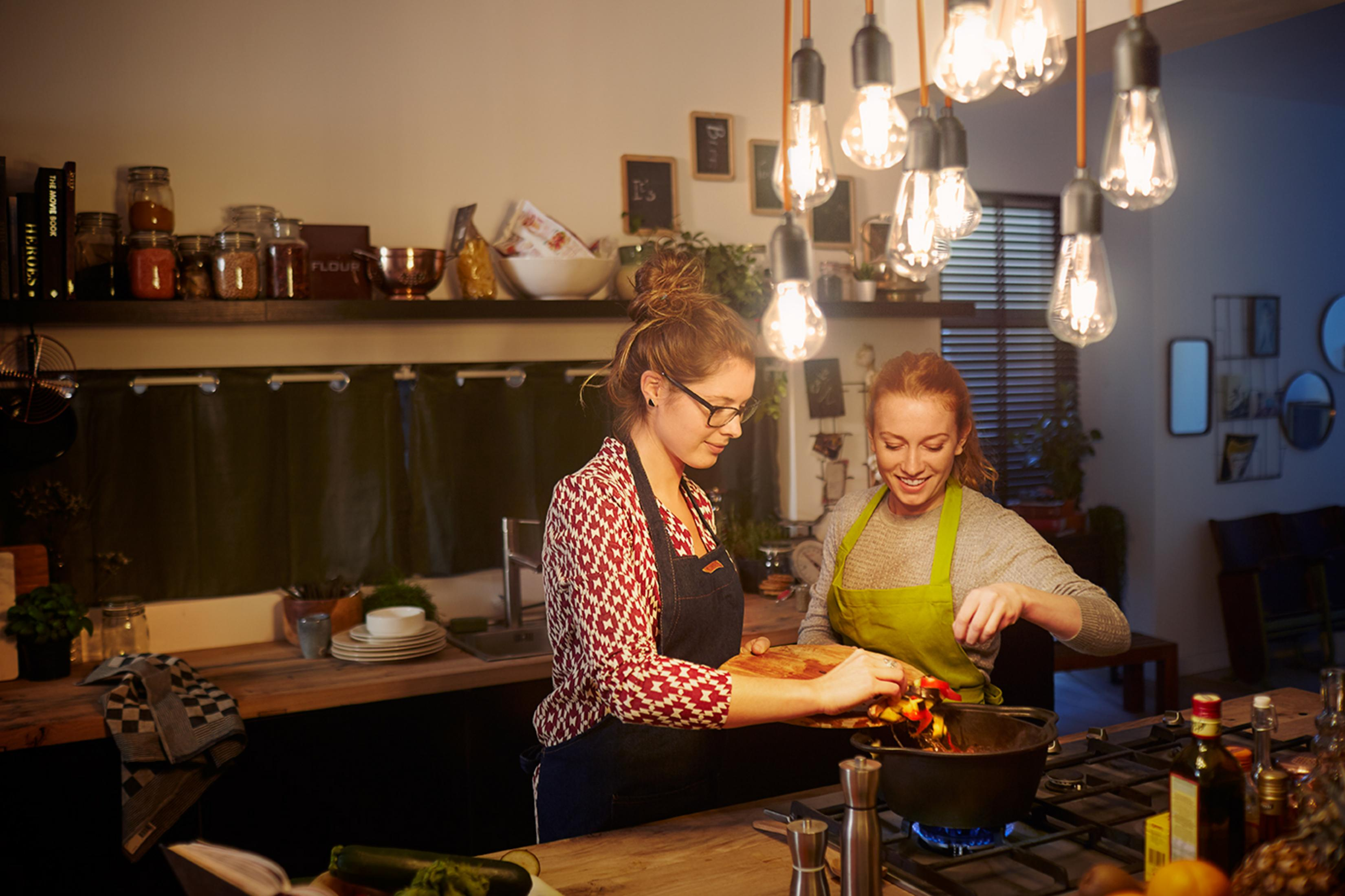Zwei Frauen in einer Küche beim Kochen mit Lichtern, die von der Decke hängen.