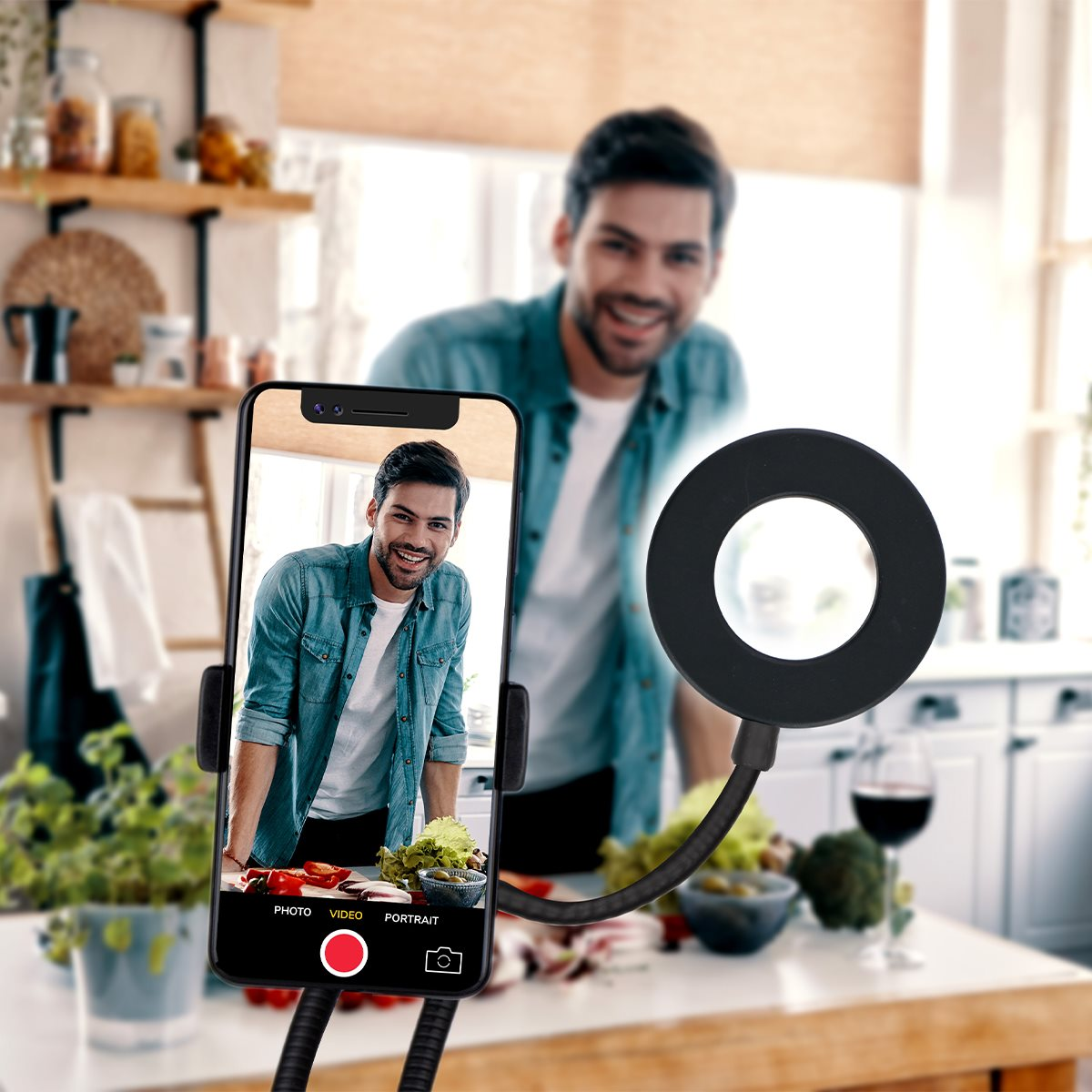 Un homme sourit, se prenant en selfie avec un téléphone et une lampe annulaire. Légumes sur la table, fond de cuisine.
