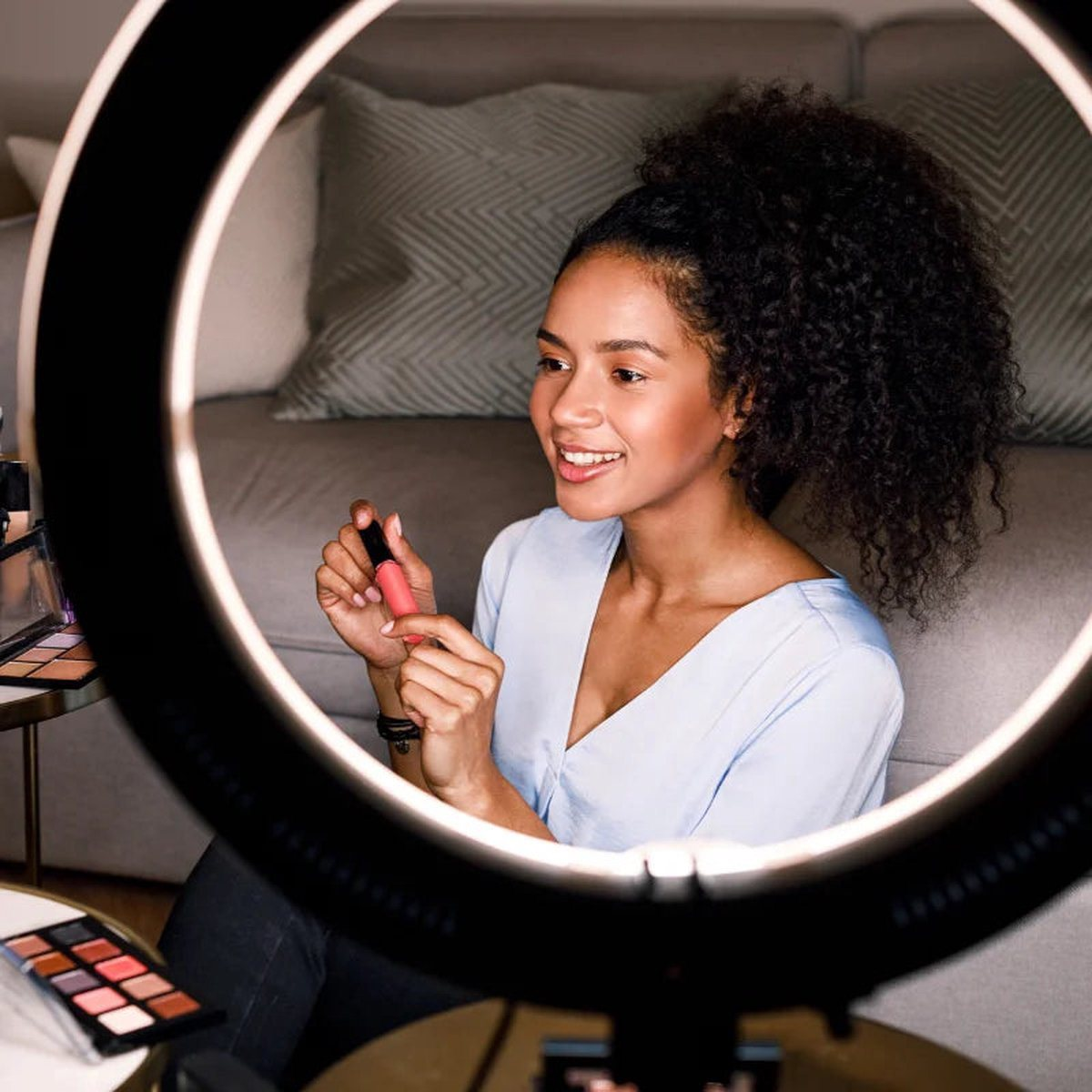 Femme appliquant du rouge à lèvres devant un anneau lumineux, maquillage sur table.