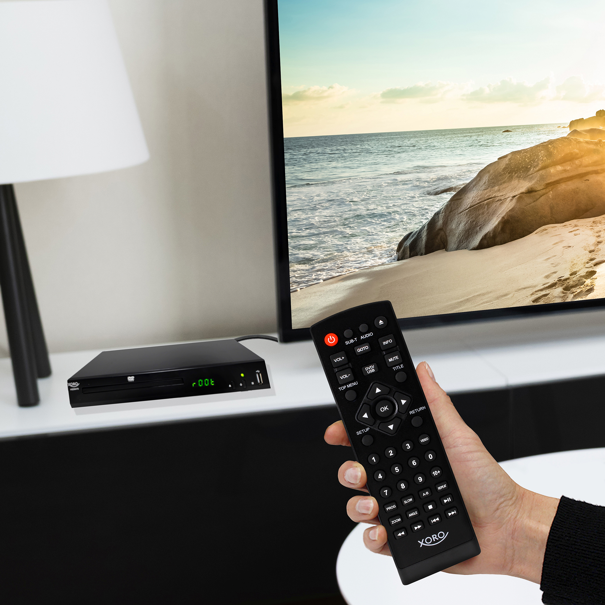 Eine Hand hält eine Fernbedienung. Ein DVD-Player steht auf einer weißen Oberfläche neben einem TV.