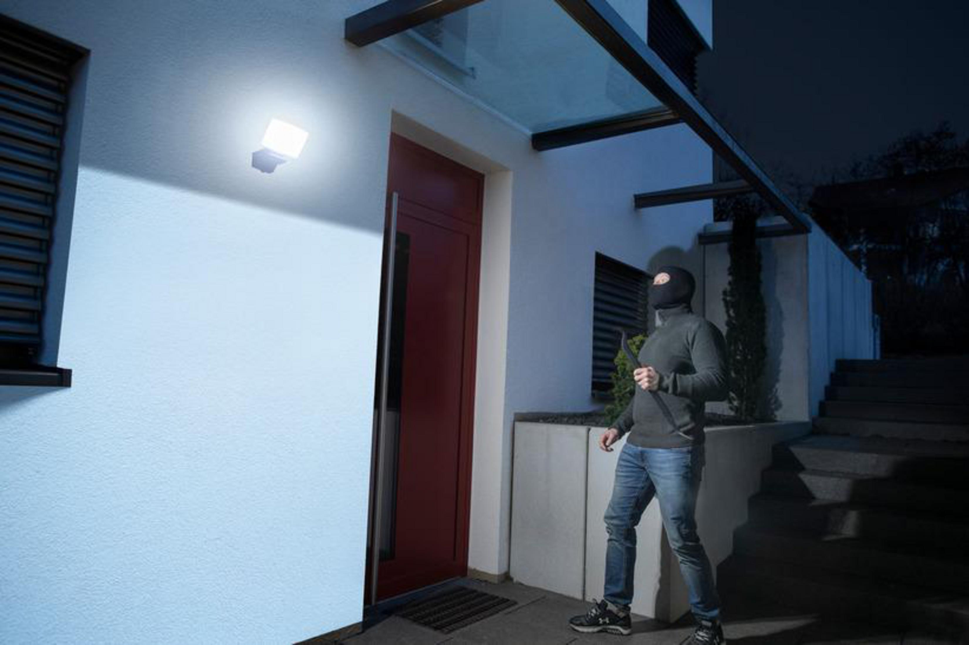 Ein Einbrecher mit Maske nähert sich einem Haus mit einem Scheinwerfer an der Wand.
