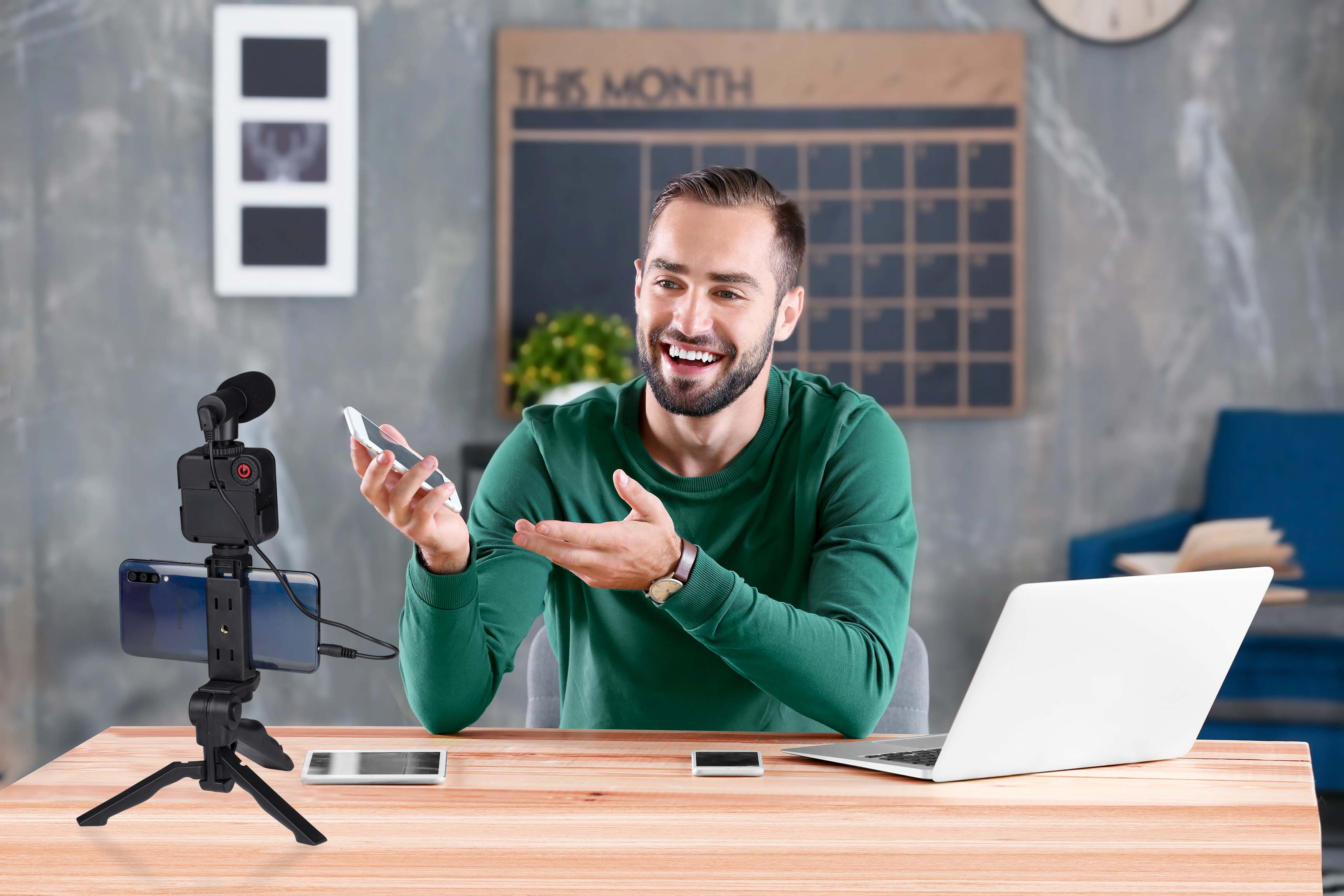 Man smiling, holding a phone, gesturing to the side. A camera setup and laptop are on the desk.