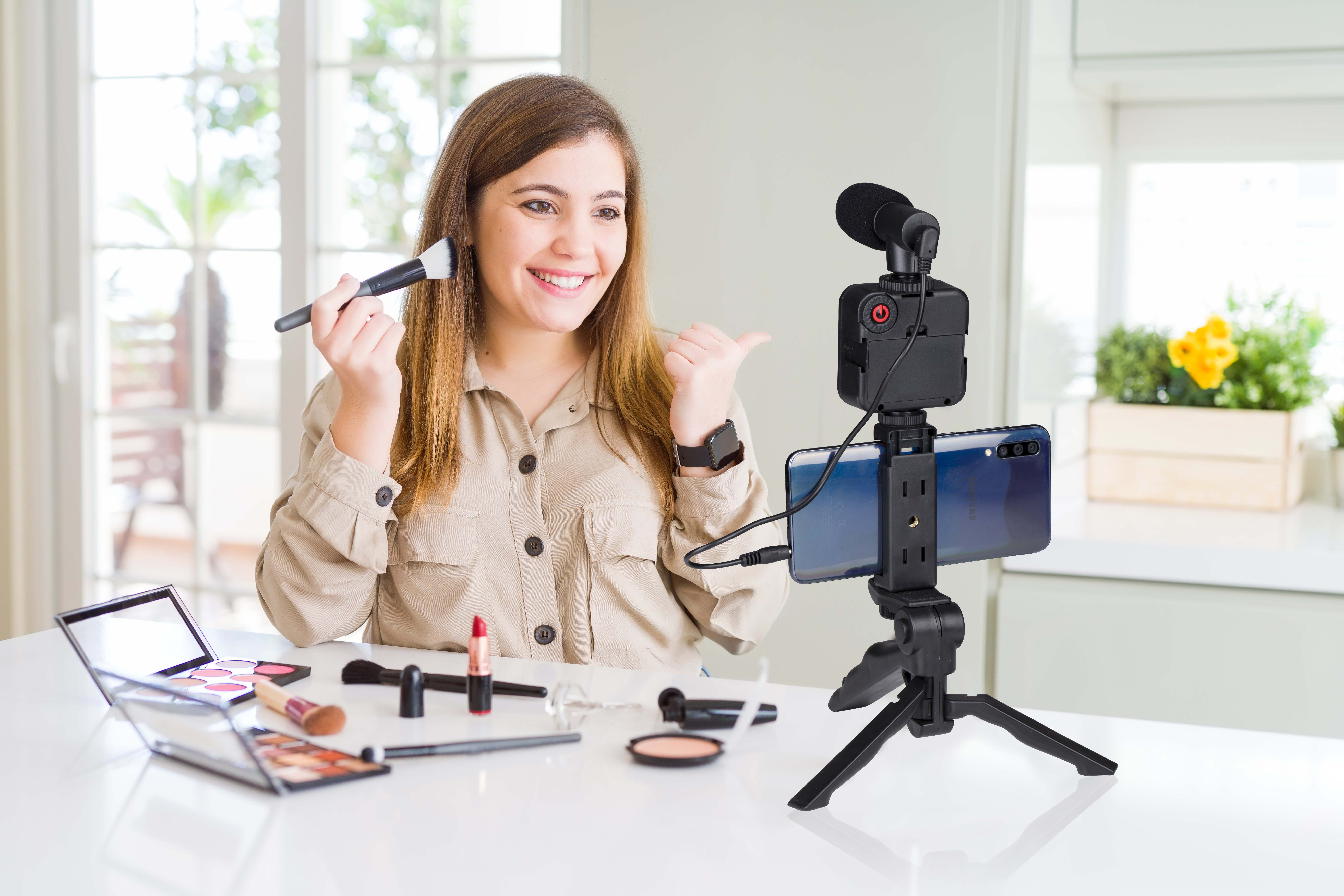 Woman applying makeup, filming with a smartphone mounted on a tripod. Makeup items are present.