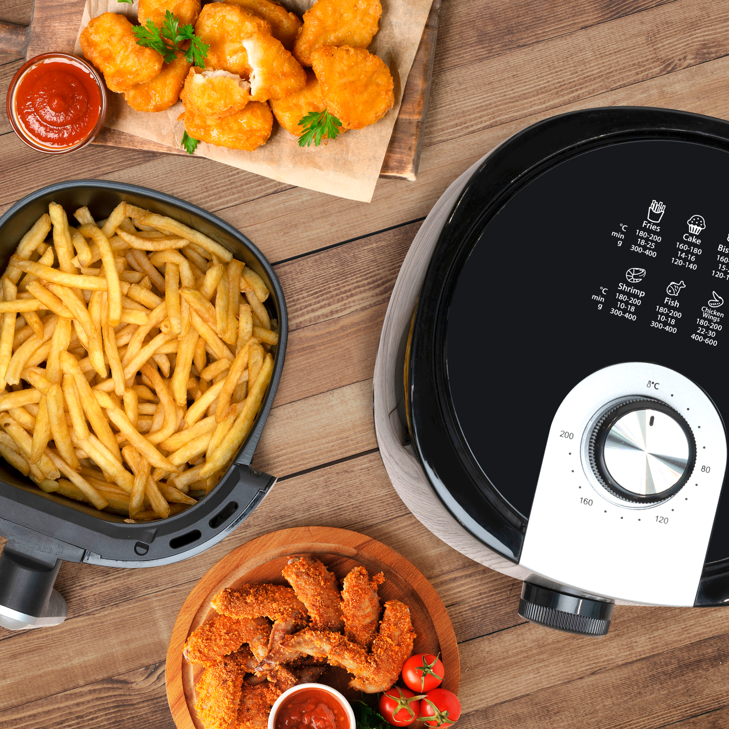 Airfryer met friet, kipnuggets en kippenvleugels op een houten tafel met sauzen.