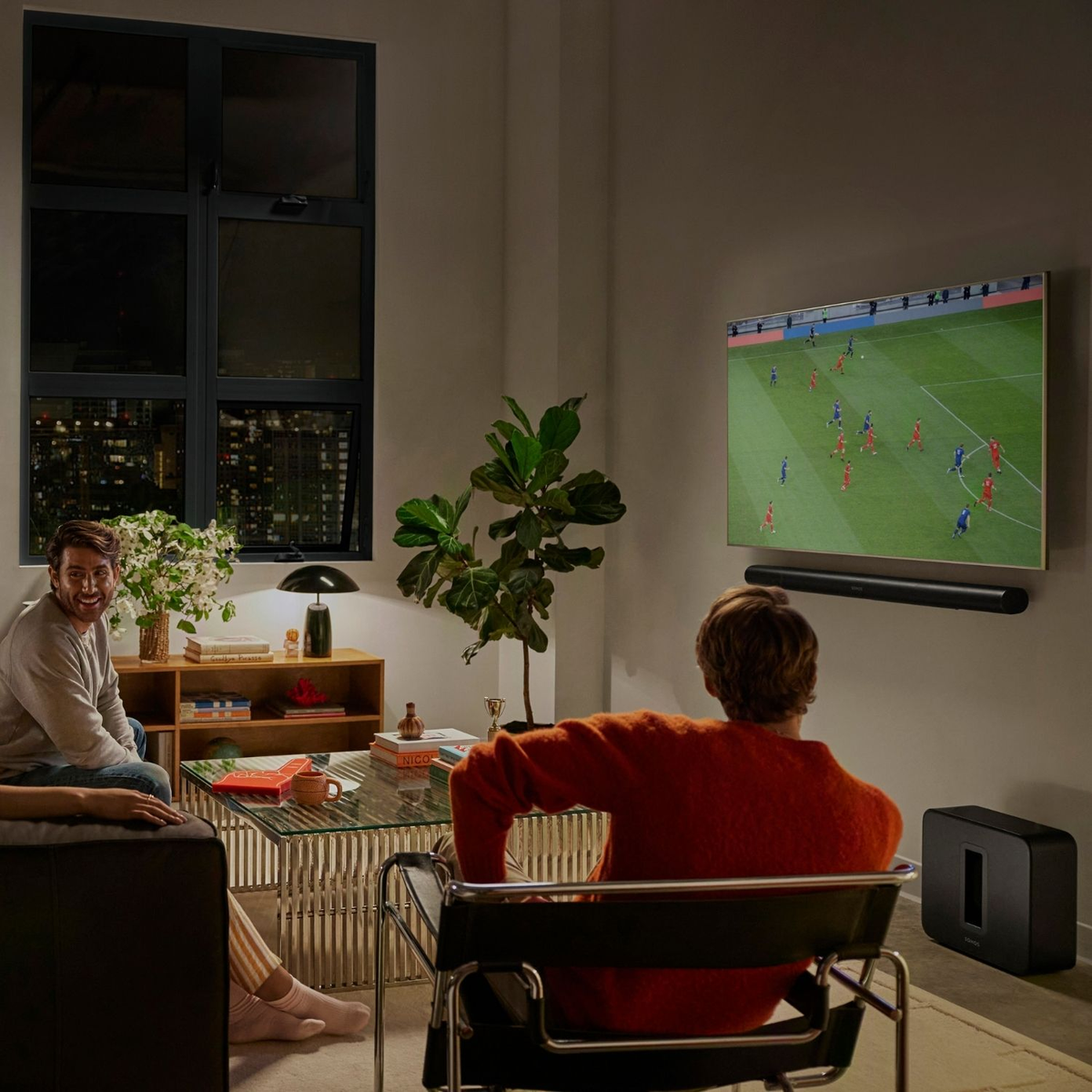 Dwóch mężczyzn ogląda piłkę nożną w telewizji. Jeden siedzi na krześle, drugi na kanapie. Zamontowano soundbar.