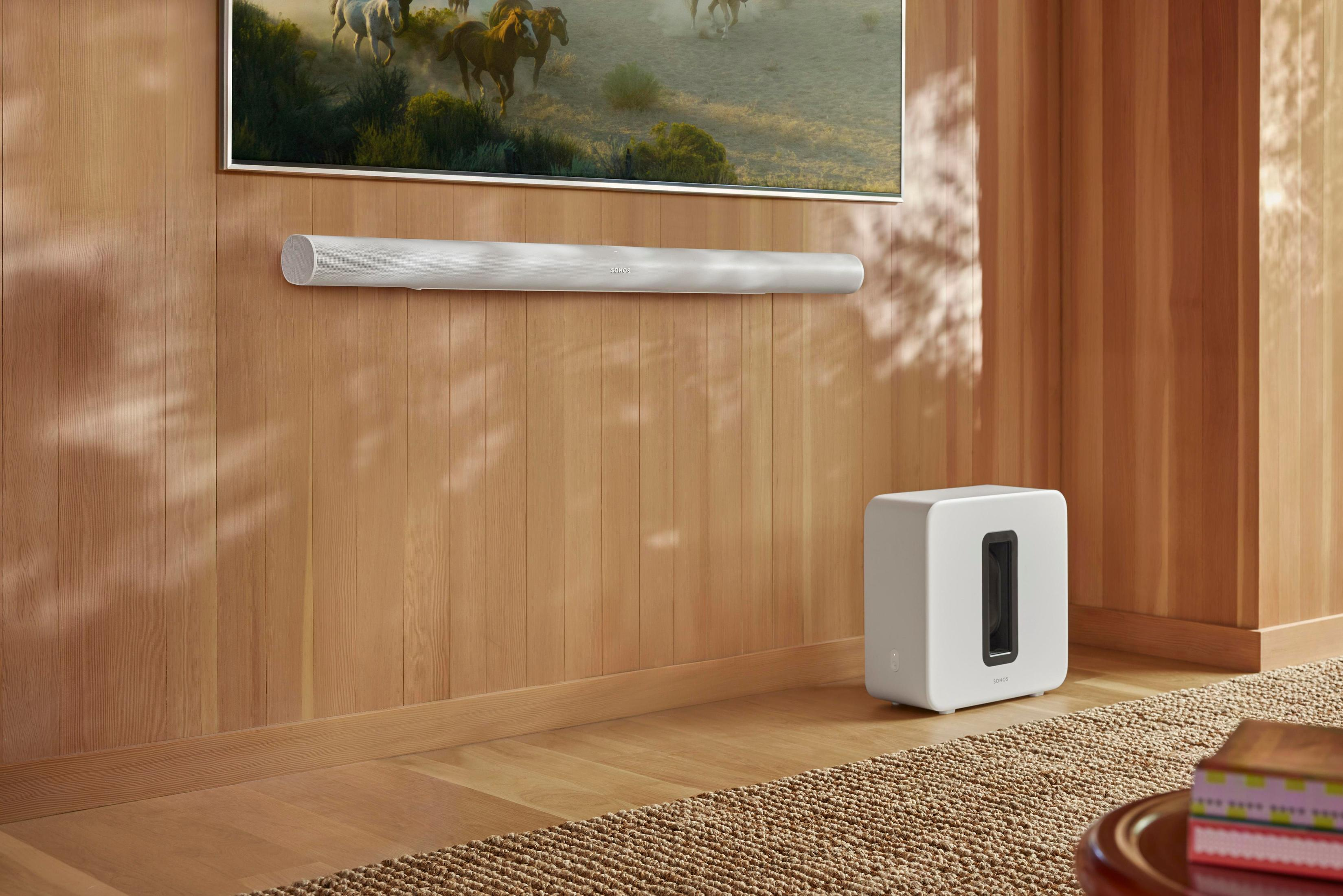 Biały soundbar i subwoofer na drewnianej ścianie, z telewizorem nad nimi. Dywan i stół z przodu.