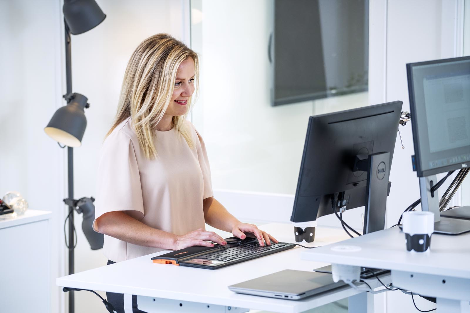 Una mujer trabajando en una computadora en un escritorio blanco, con dos monitores y un teclado.