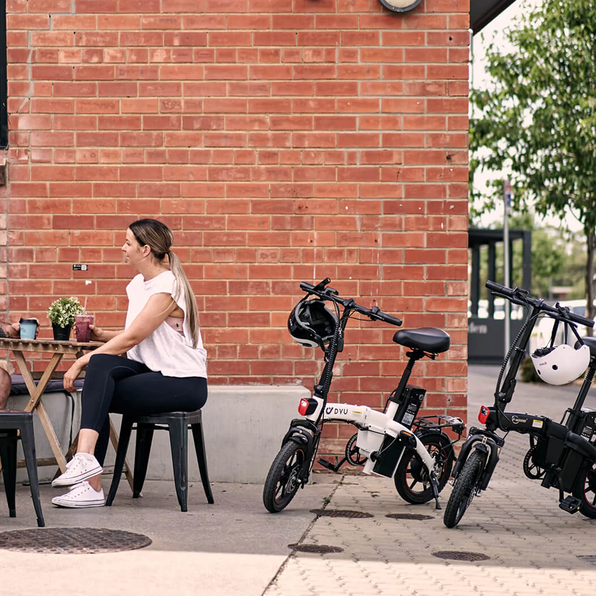 Kobieta siedzi przy stole w pobliżu dwóch rowerów elektrycznych zaparkowanych na chodniku, na tle ceglanej ściany.