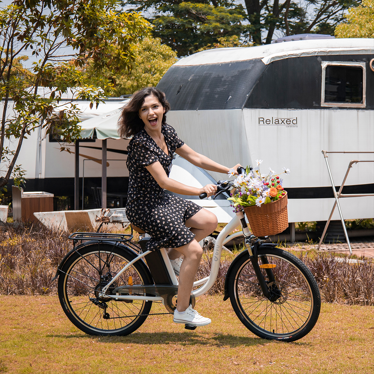 Kobieta jedzie na rowerze z koszem kwiatów. W tle kamper i drzewa.