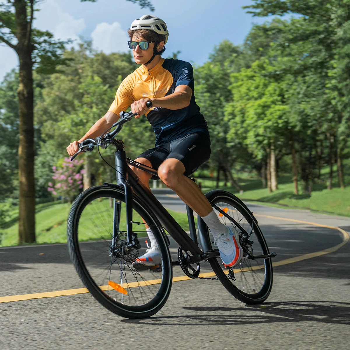 Mężczyzna jadący czarnym rowerem po utwardzonej drodze, otoczony drzewami. Ma na sobie okulary przeciwsłoneczne i kask.