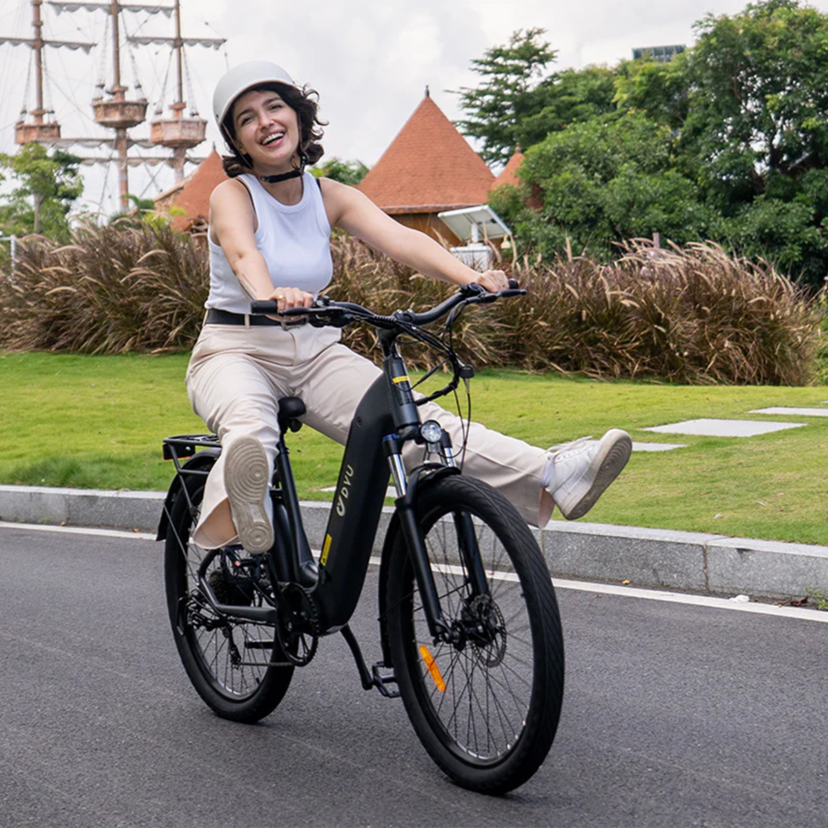 Kobieta jedzie czarnym rowerem elektrycznym, uśmiechnięta, w białym kasku na utwardzonej ścieżce.