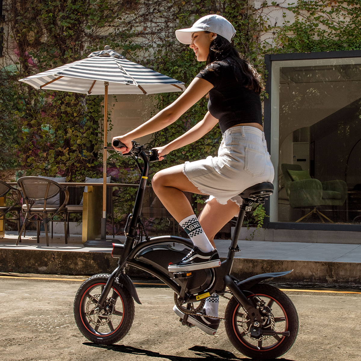 Kobieta jadąca na czarnym rowerze elektrycznym, w białej czapce i szortach, w słoneczny dzień.