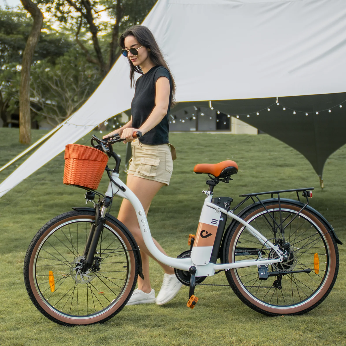 Kobieta w okularach przeciwsłonecznych idzie obok białego i pomarańczowego roweru elektrycznego na trawie, z białym namiotem z tyłu.
