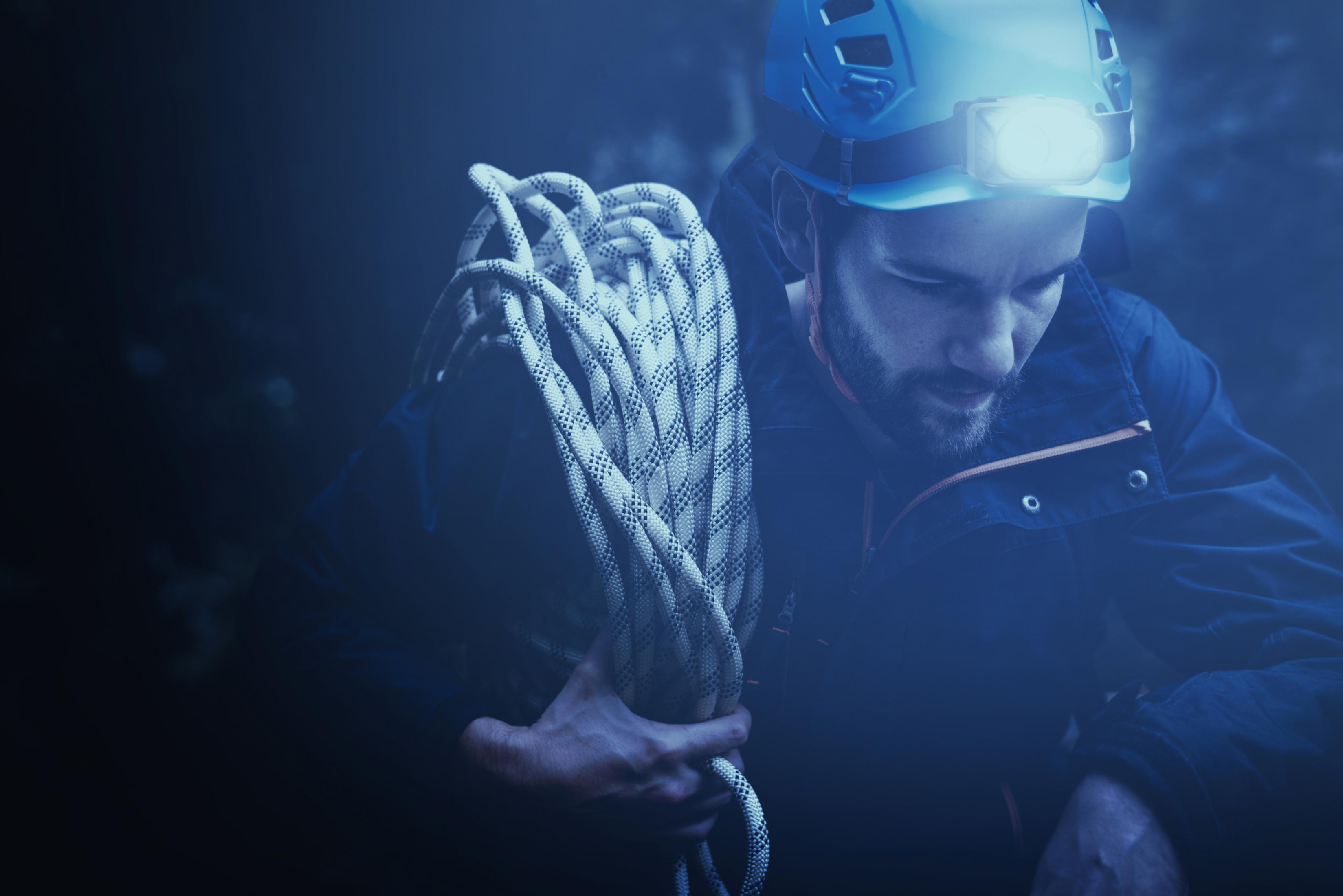 Mężczyzna w kasku i czołówce trzyma zwiniętą linę. Scena jest ciemna i na zewnątrz.